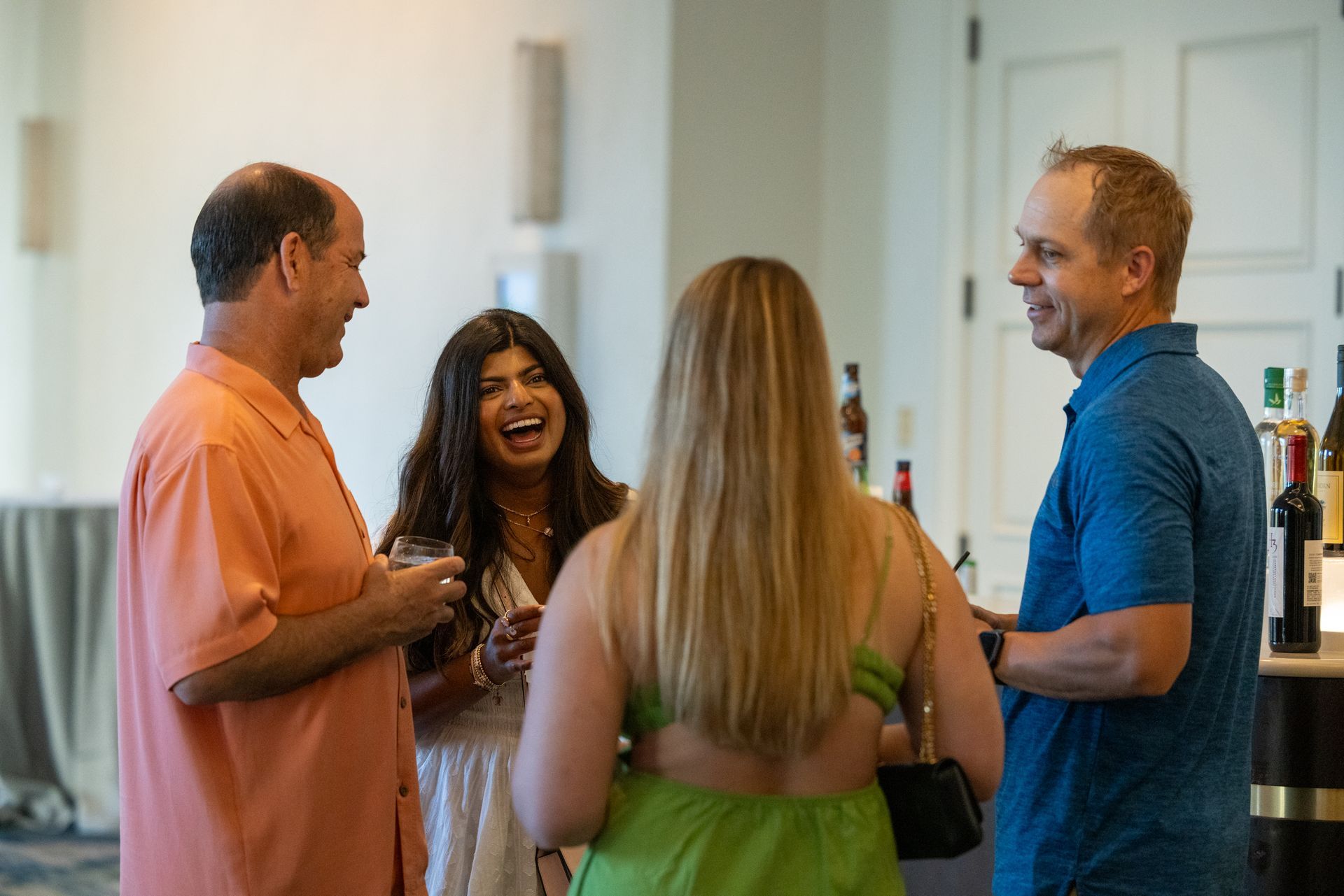 Four people socializing, laughing, holding glasses near a wine display.