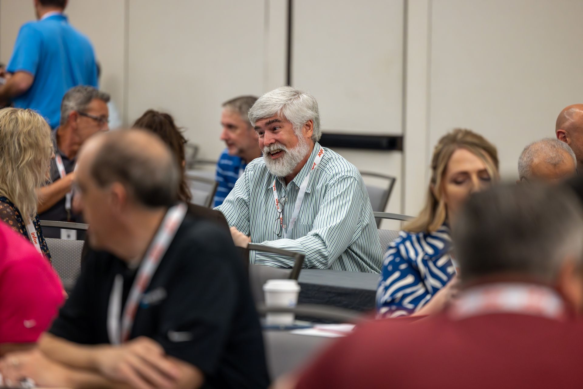 Man with a gray beard talking at a conference table, wearing a striped shirt. Others are seated nearby.