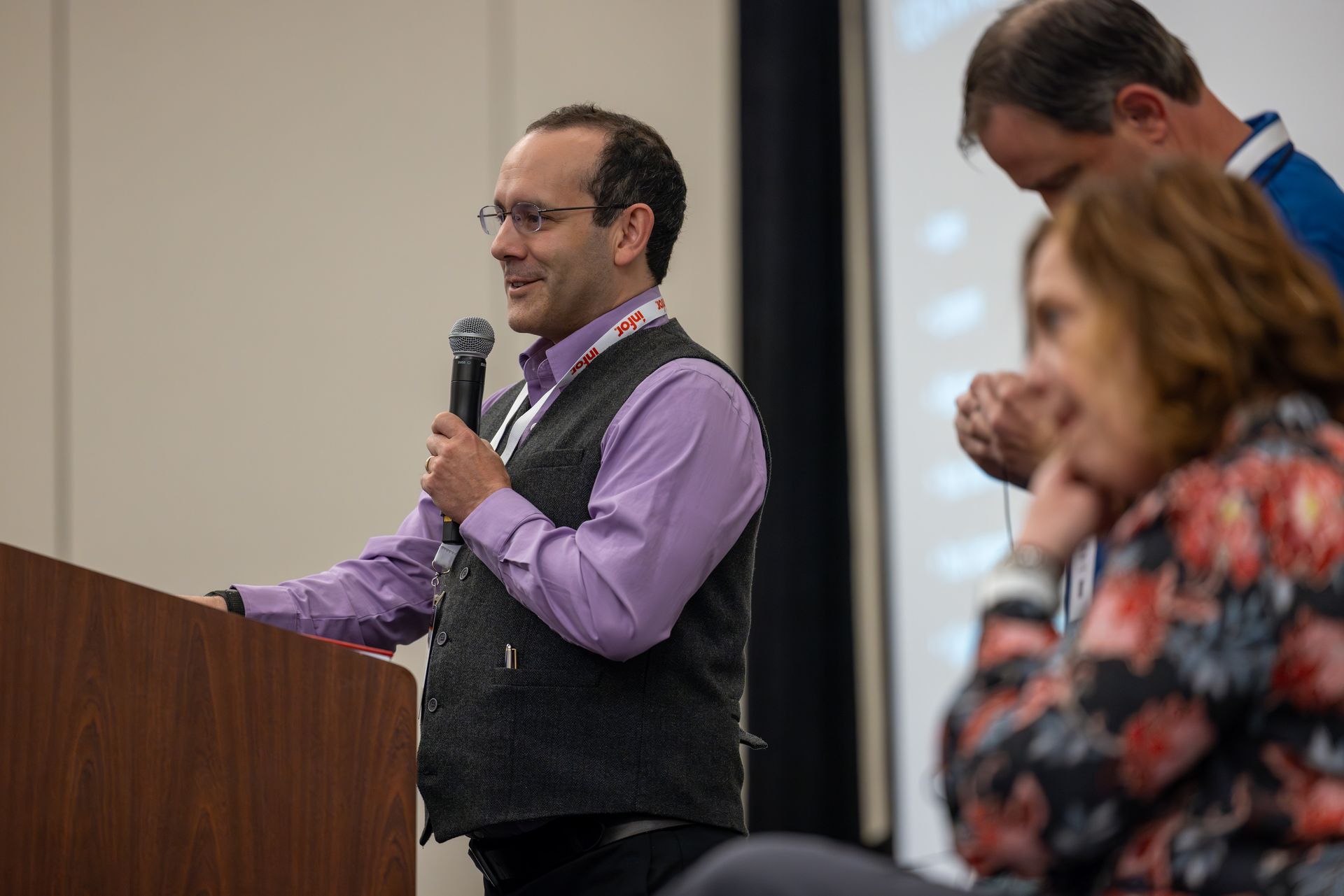 Man in purple shirt, vest, speaking into a microphone at a podium. People in background.