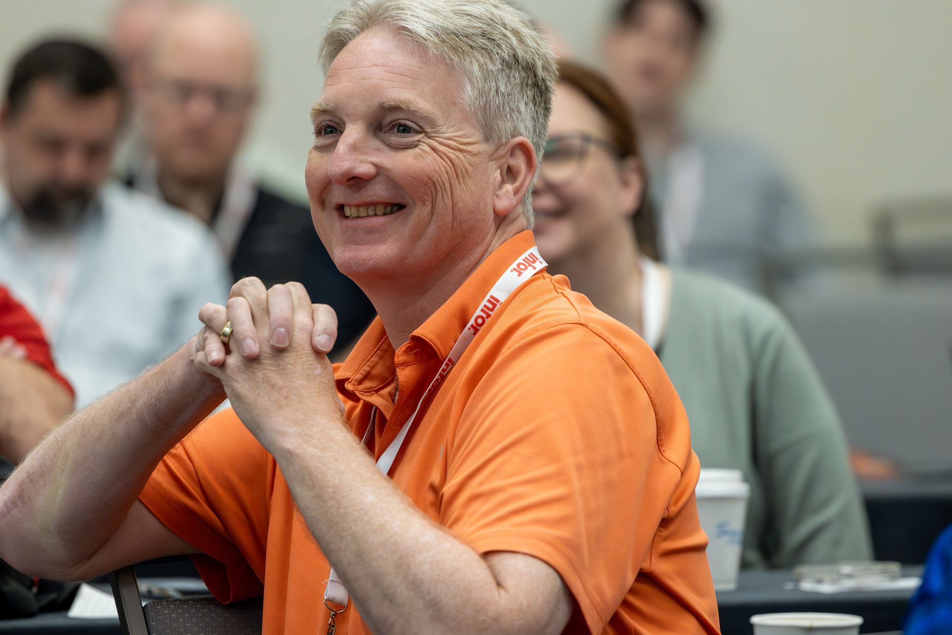 Man in orange shirt smiles, hands clasped, in a conference setting. Other attendees are blurred in the background.