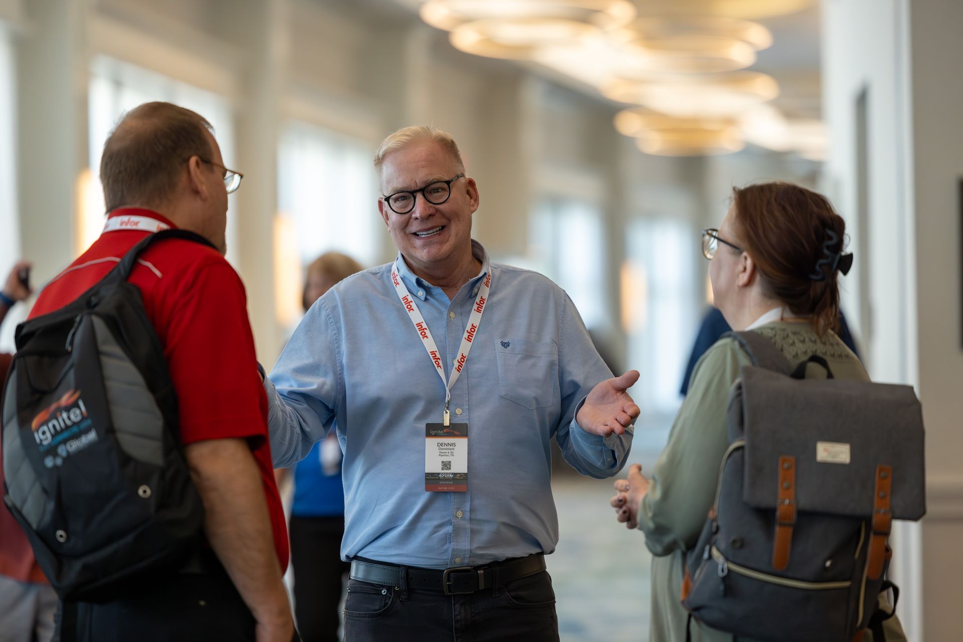 Three people conversing in a brightly lit hallway. Man in center gestures, smiling, wearing glasses and blue shirt.