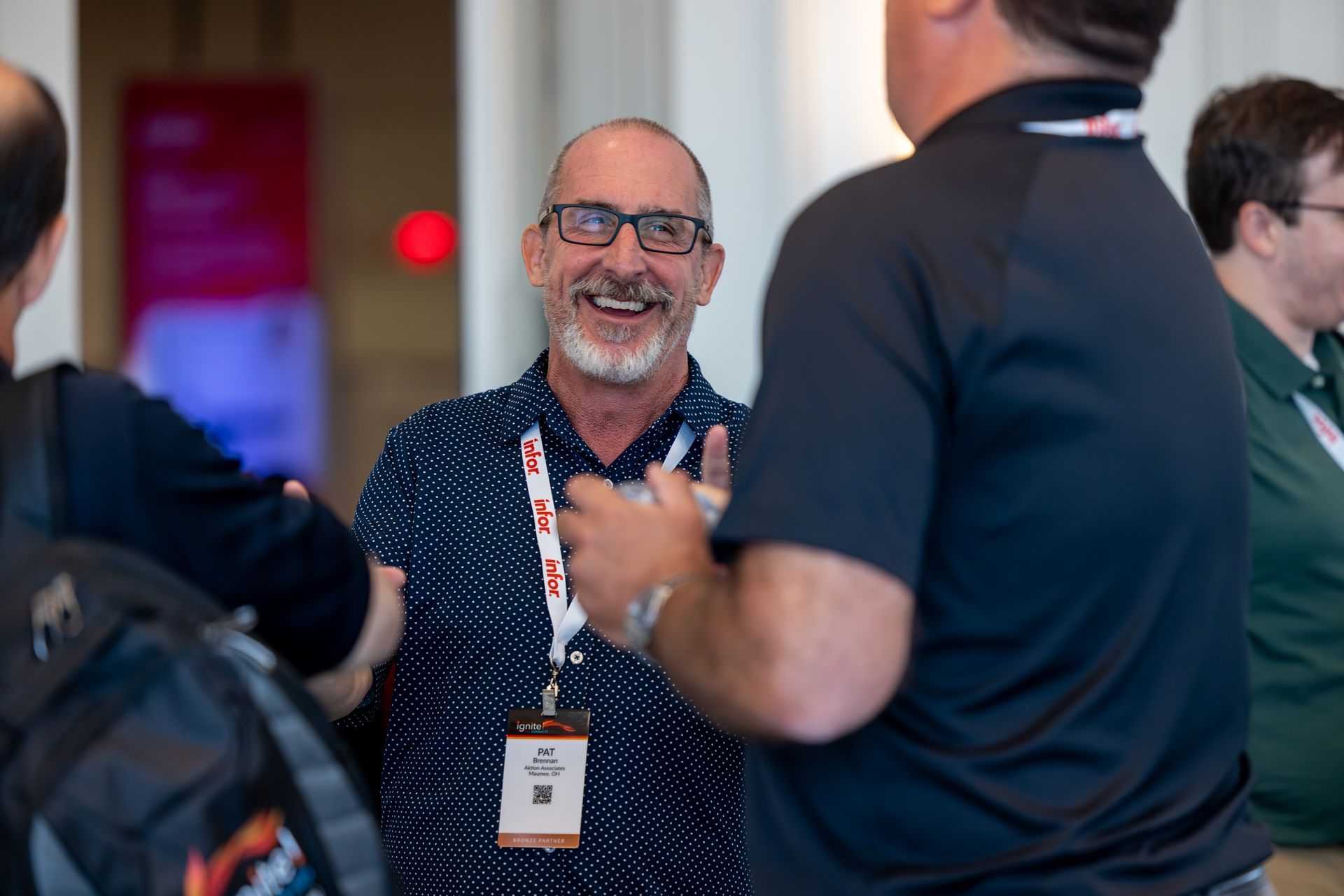 Man with glasses smiles, talking with others at conference. He wears patterned shirt, lanyard.
