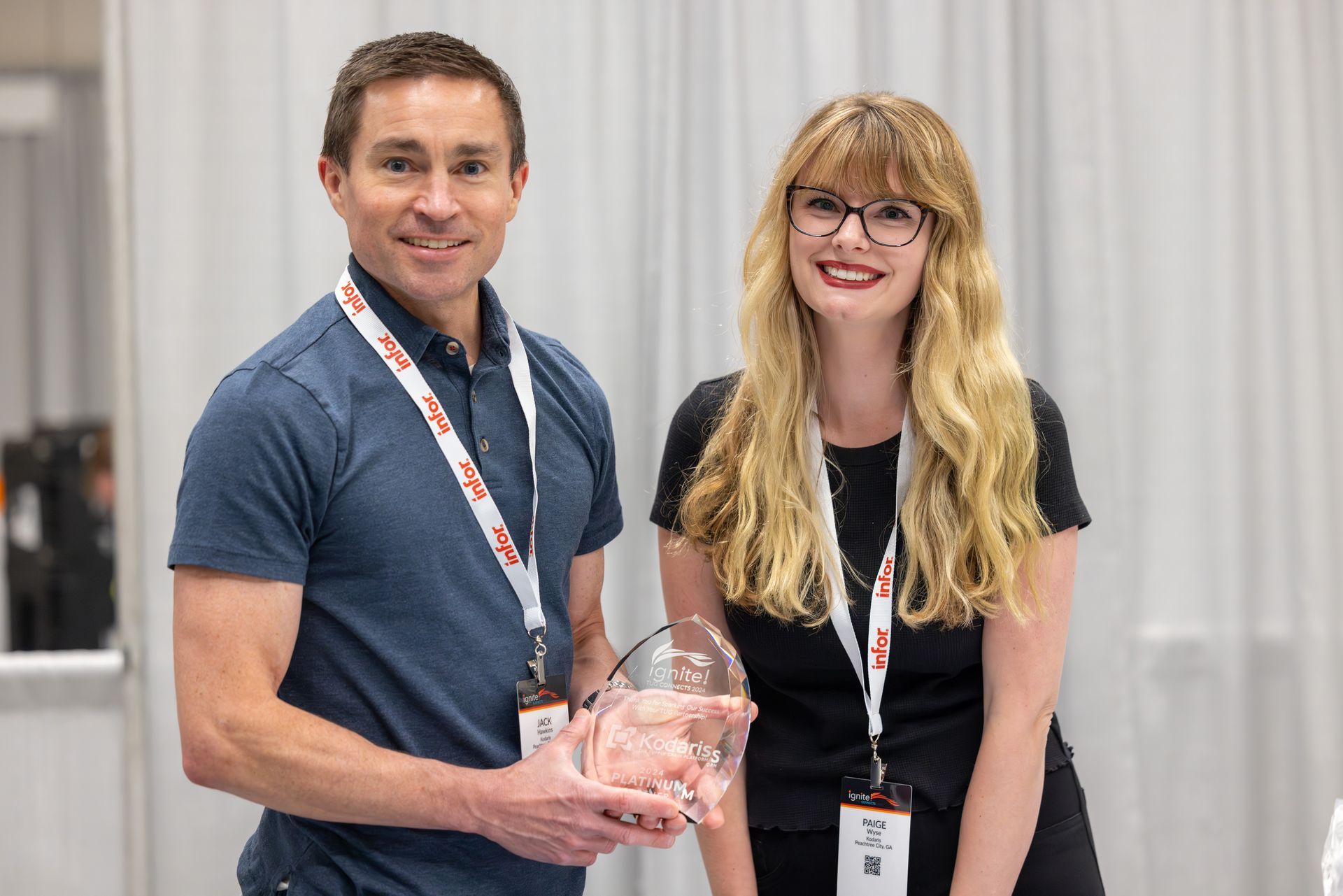 Man and woman holding an award, smiling at a conference. They wear lanyards, and stand in a white space.