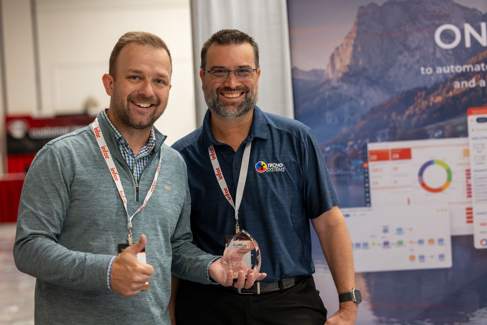 Two men smile, one holds a trophy. Behind them is a display with graphics and the word 