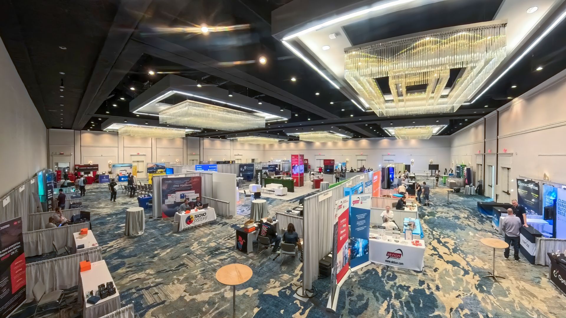 Large event hall with vendor booths and attendees. Blue carpet, bright lights, multiple chandeliers.