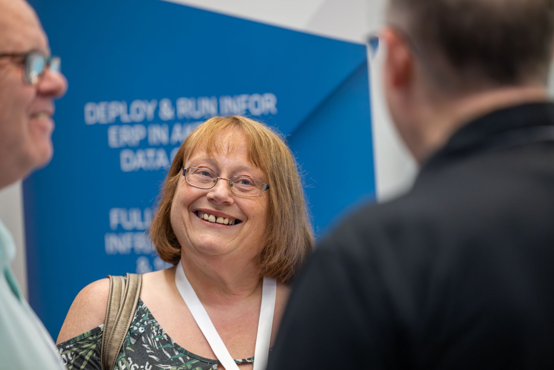 Woman with glasses smiles while talking to two men at a conference; a blue banner is in the background.