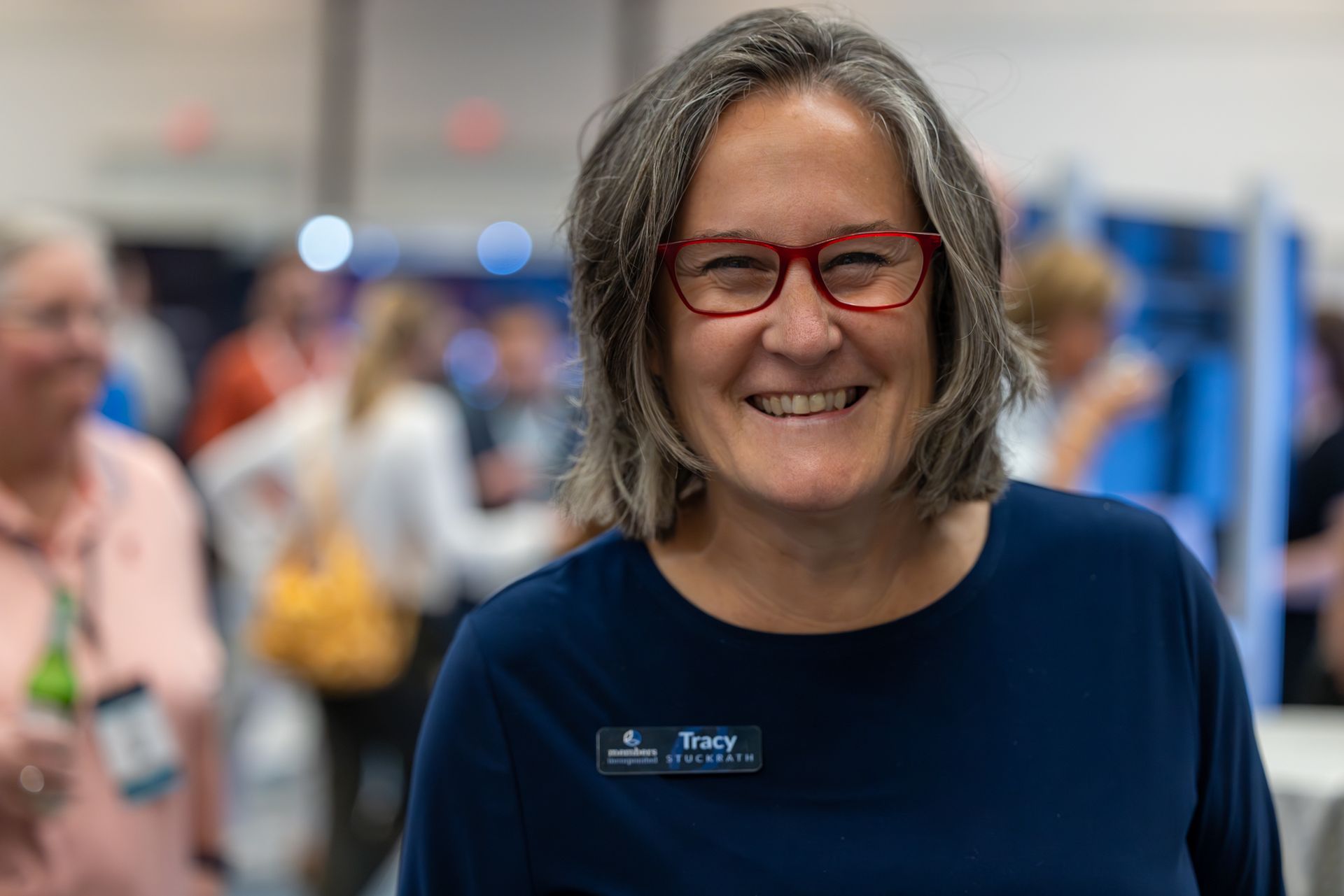 Woman with red glasses smiles at the camera, wearing a blue shirt with a name tag, at a conference.