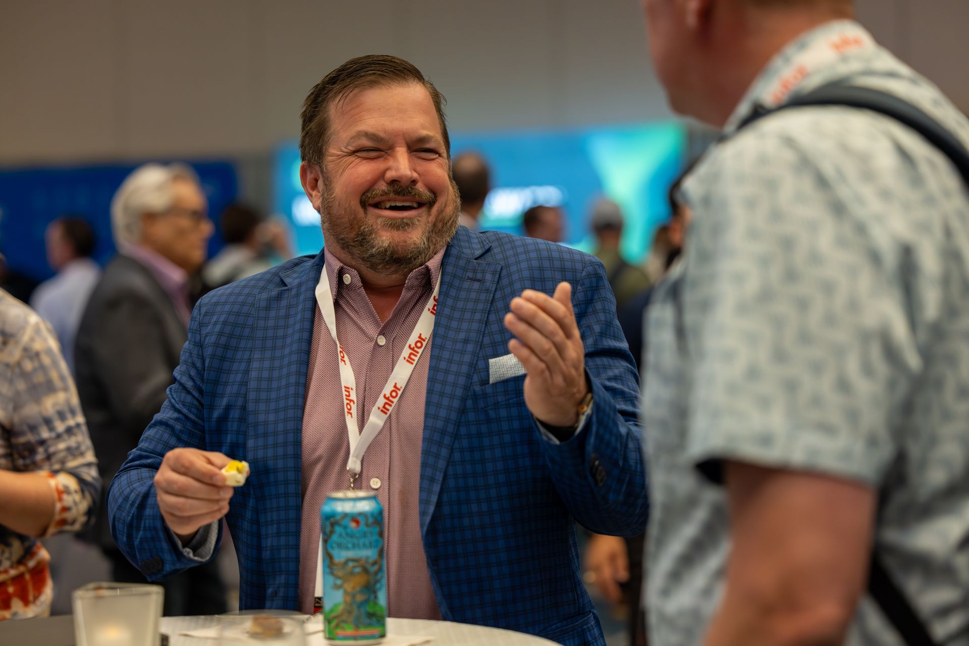 Man in blue blazer laughs, holding a drink, talking to another person at an event.