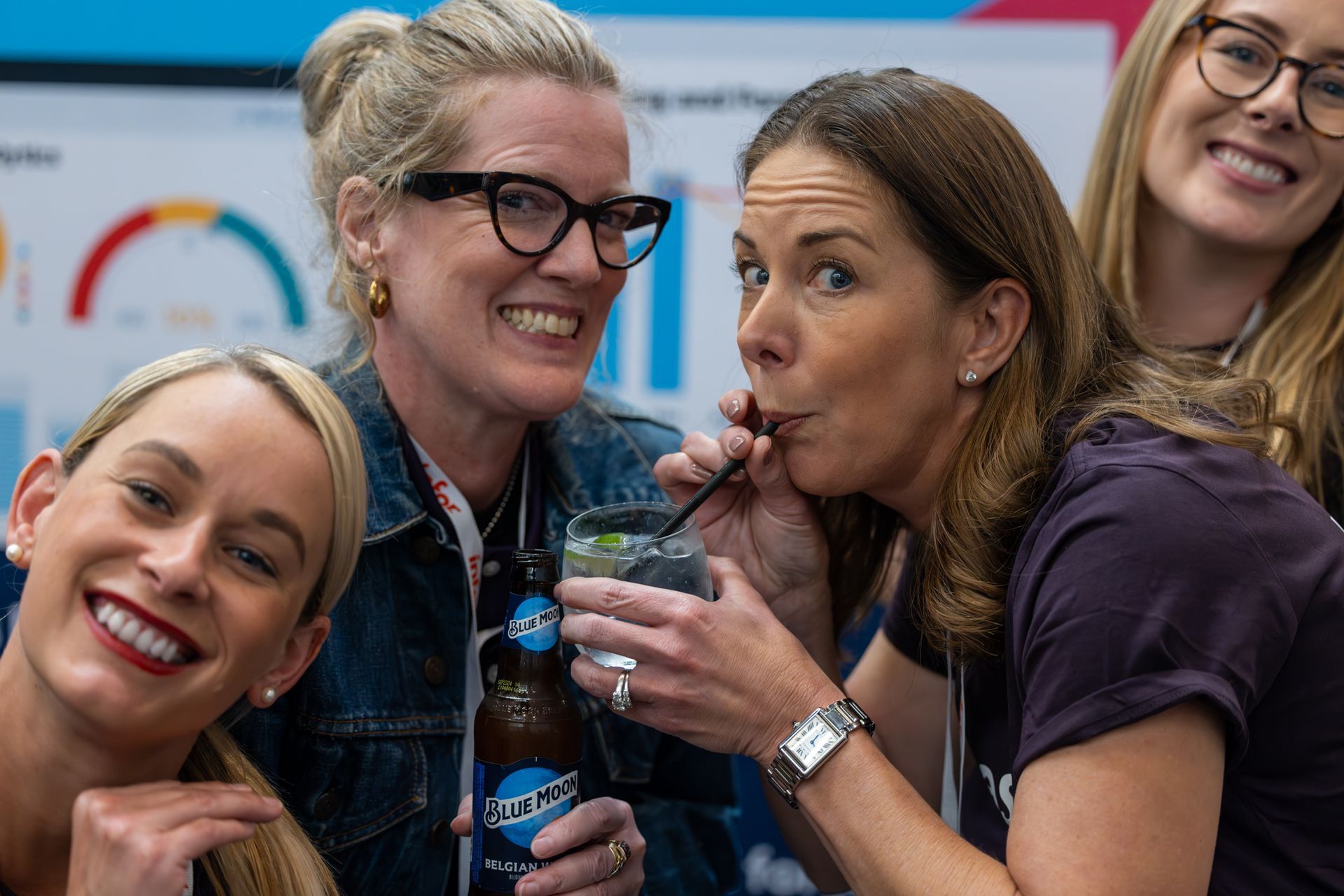 Four women smiling, one drinking from a straw, holding a beverage with a logo on the bottle, in front of a colorful chart.