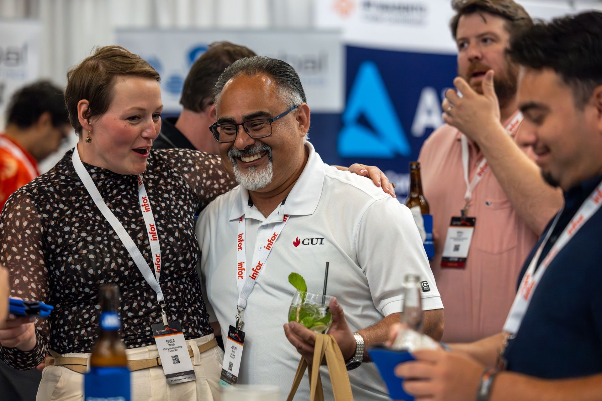 People at an event laughing and interacting. Man in white polo holds a cocktail. Others hold drinks and badges.