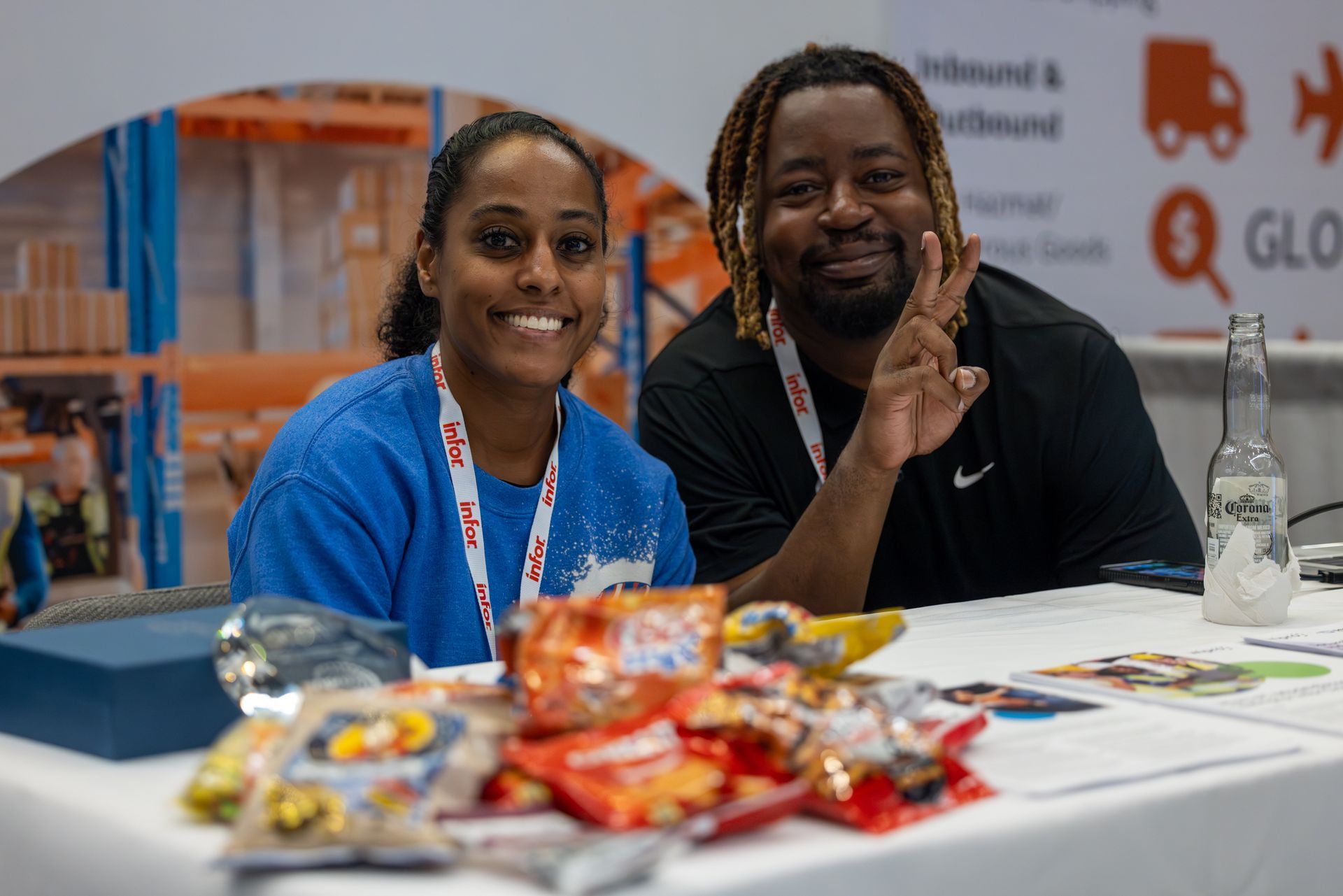 Two people at a table smiling, snacks in front. A backdrop displays logistics symbols.