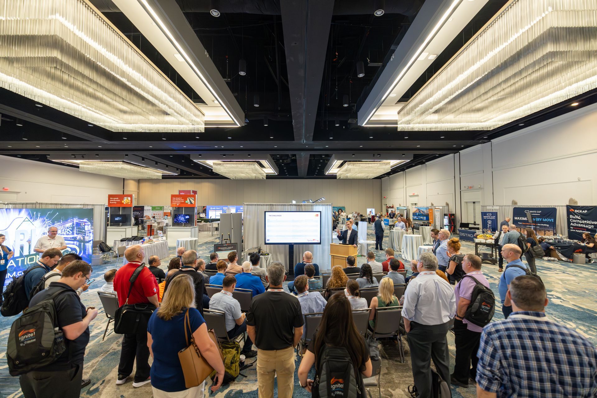 Conference attendees gather in a large room, watching a presentation on a screen.