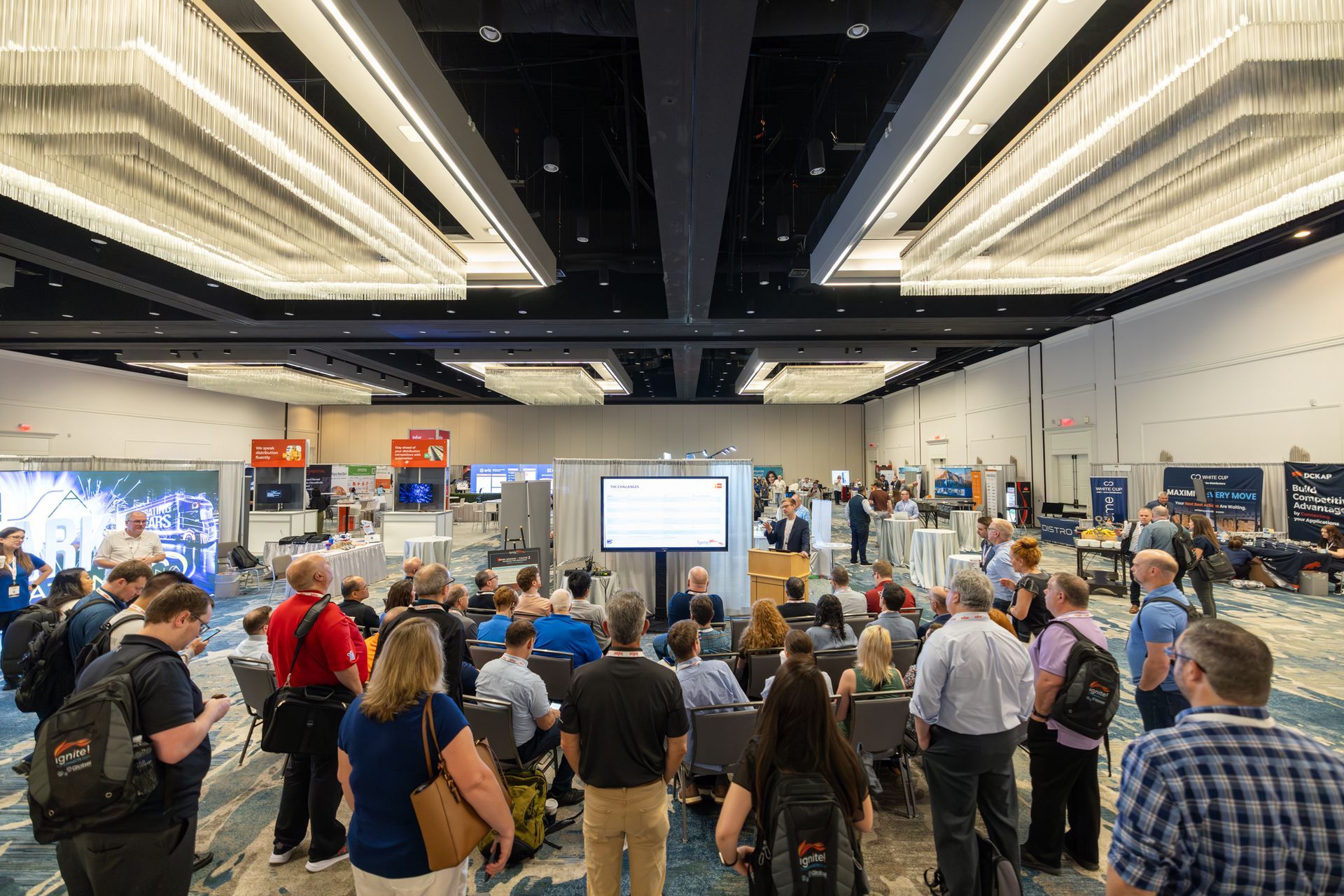 Conference room with a presentation in progress; attendees seated and standing, looking at the speaker.