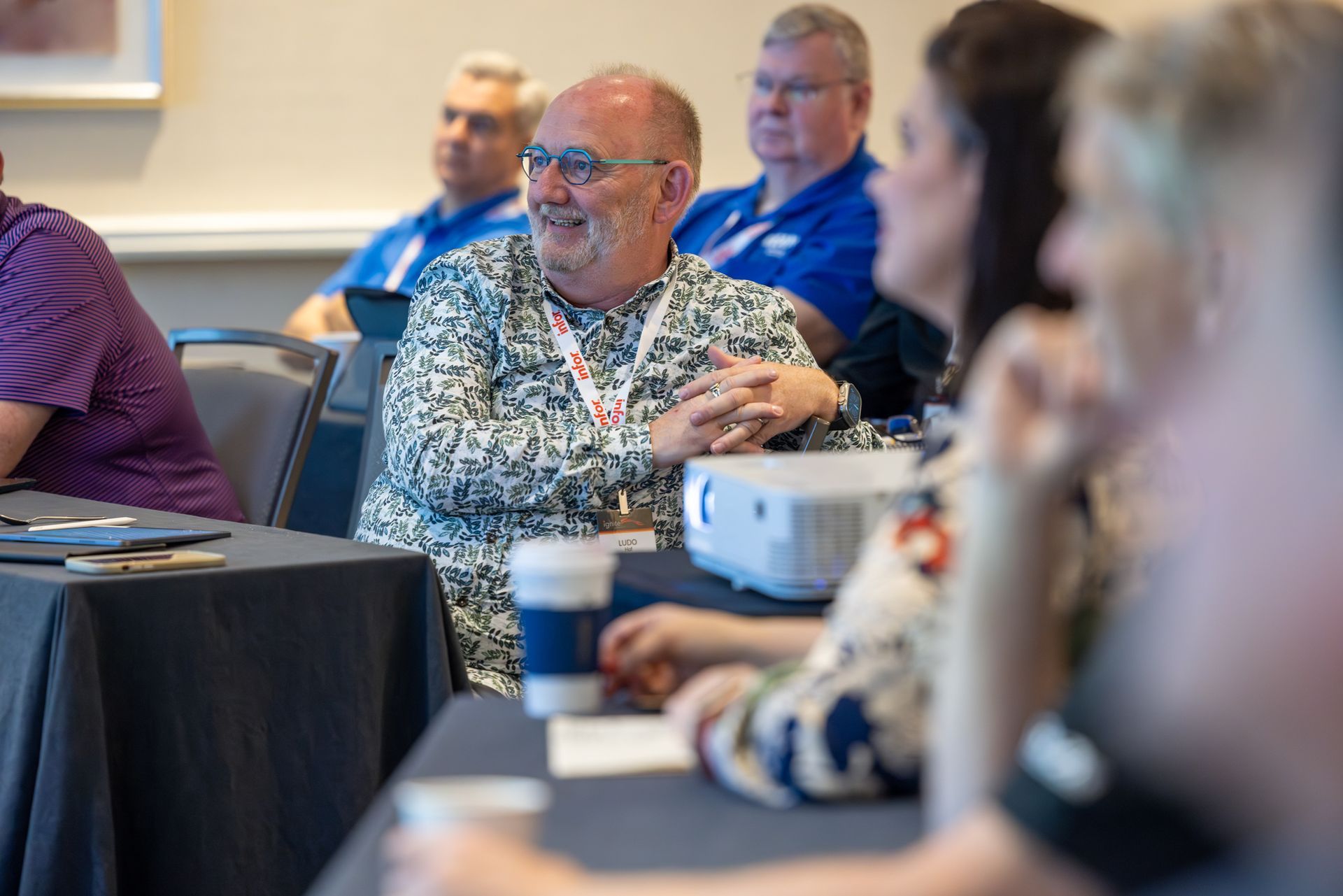 Man in patterned shirt smiles at a conference, seated at a table. Others listen in the background.