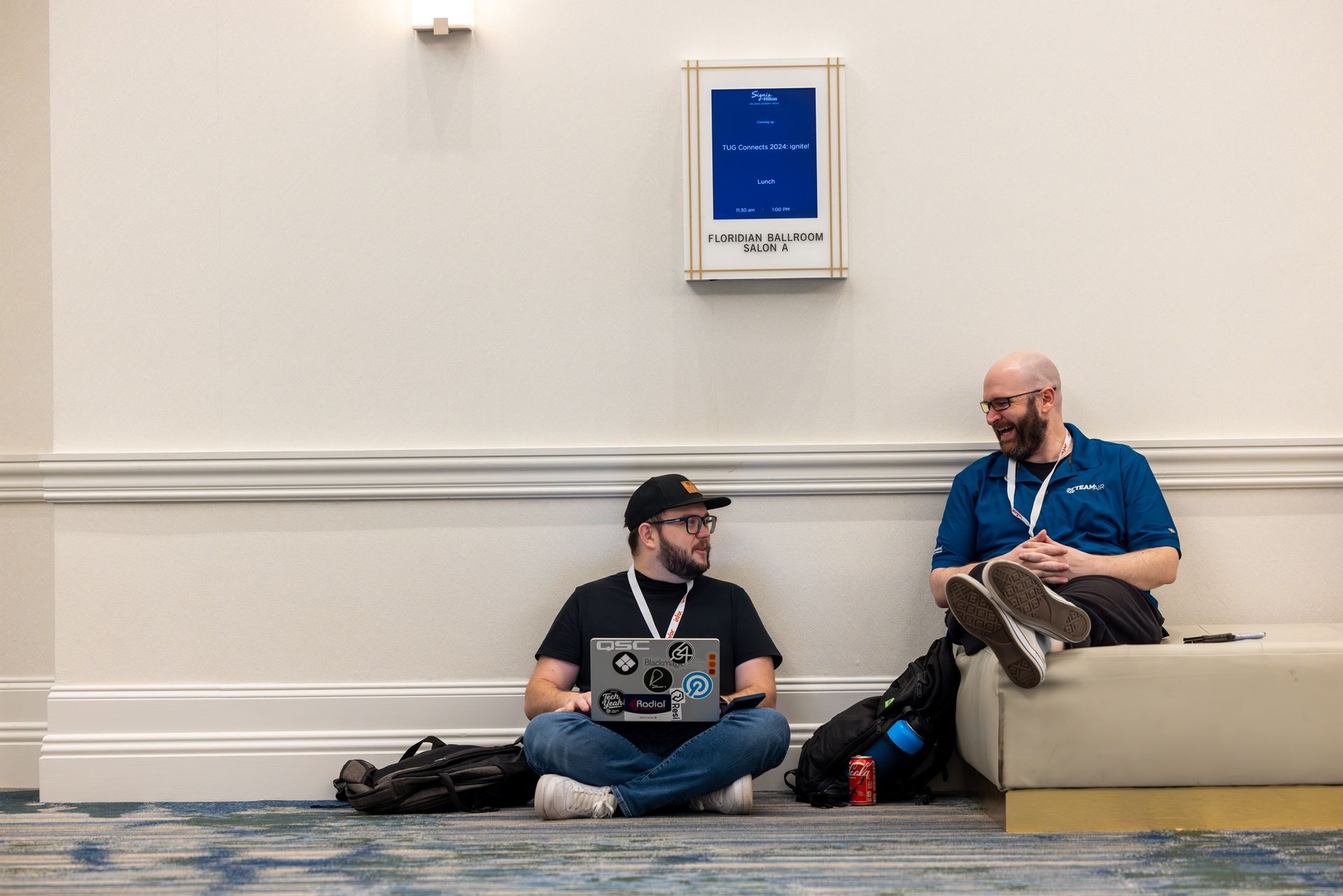 Two men seated indoors, one using a laptop, the other resting on a bench, talking.