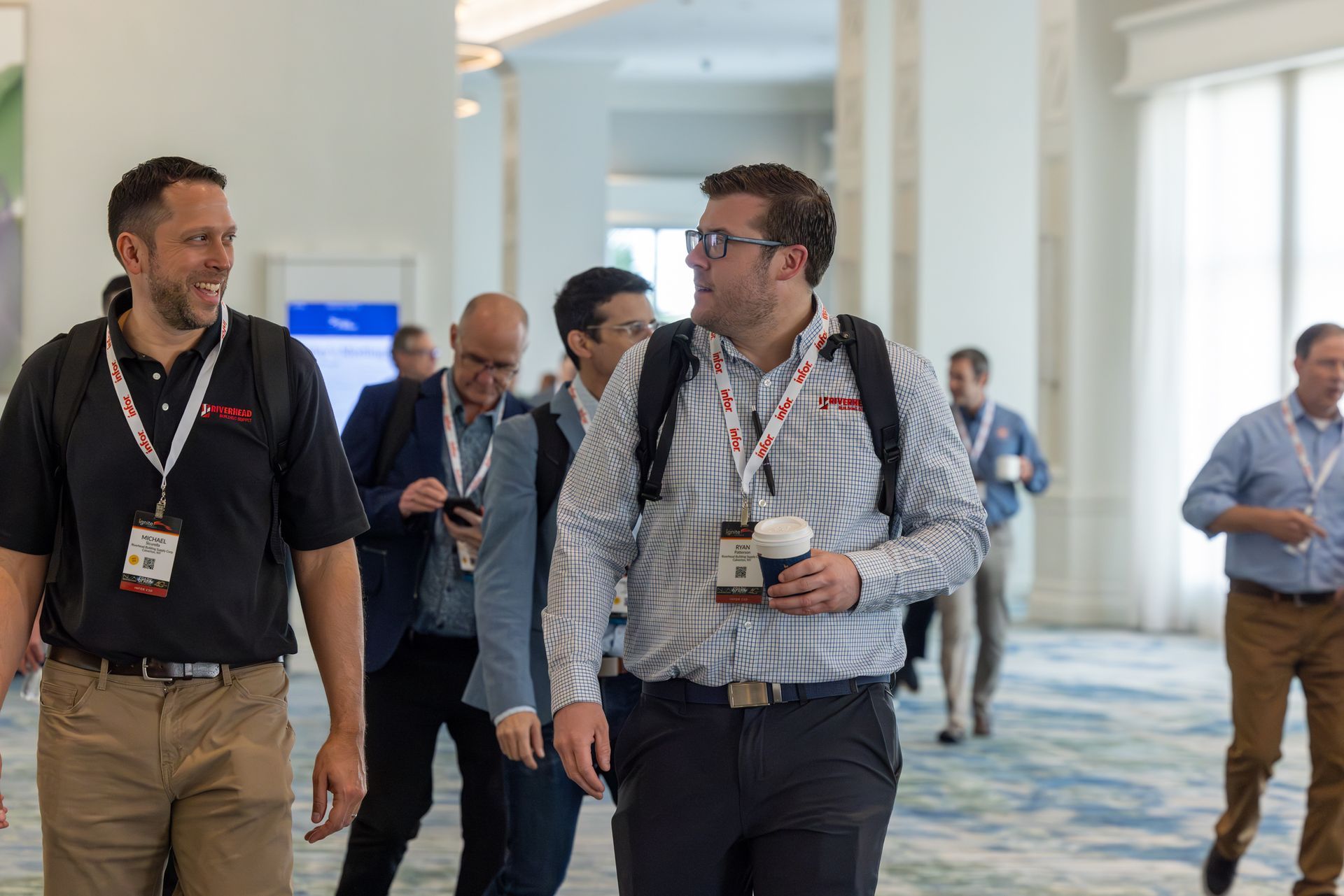 People walking in a conference hallway, some wearing lanyards and carrying coffee, talking and smiling.
