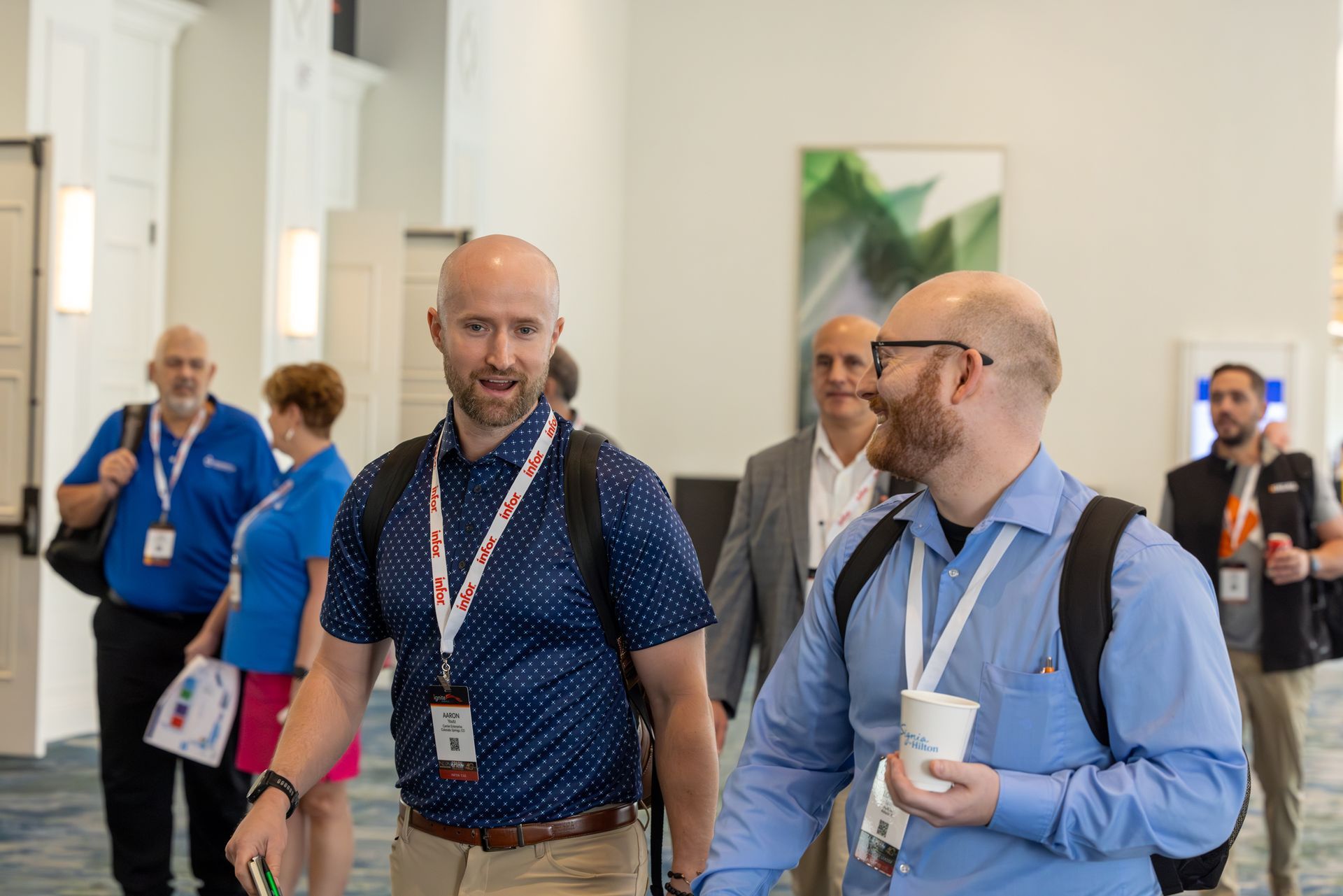 People walking in a conference hallway; two bald men wearing lanyards and backpacks converse.