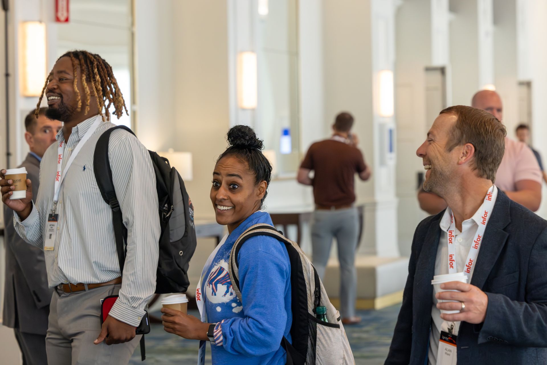 People smiling and walking in a bright hallway, some carrying coffee cups and backpacks, likely at a conference.