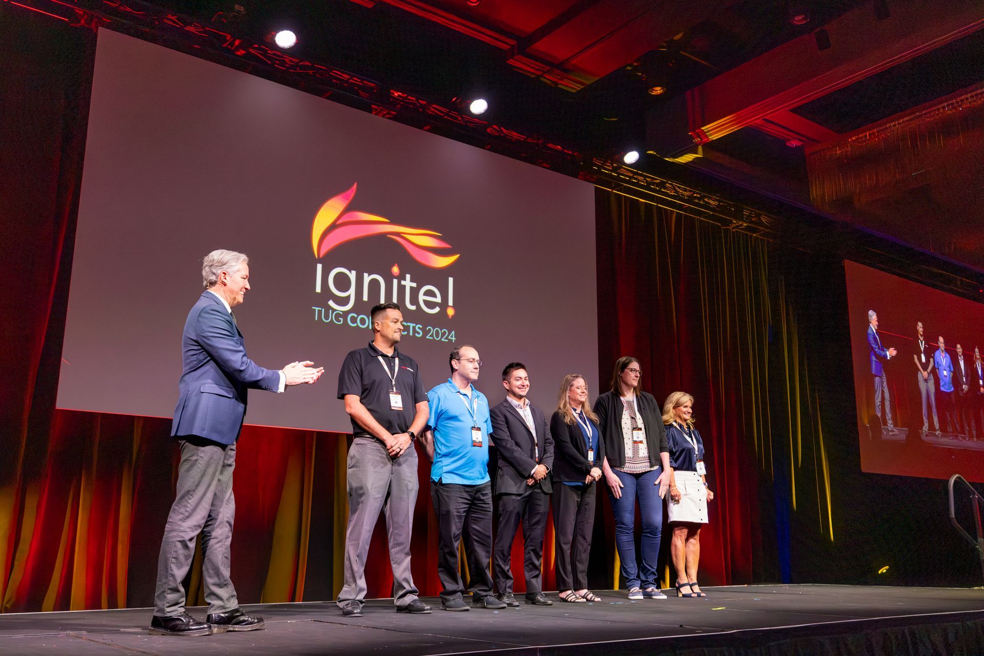 Man on stage with a group, event logo backdrop; a conference setting with bright lights.