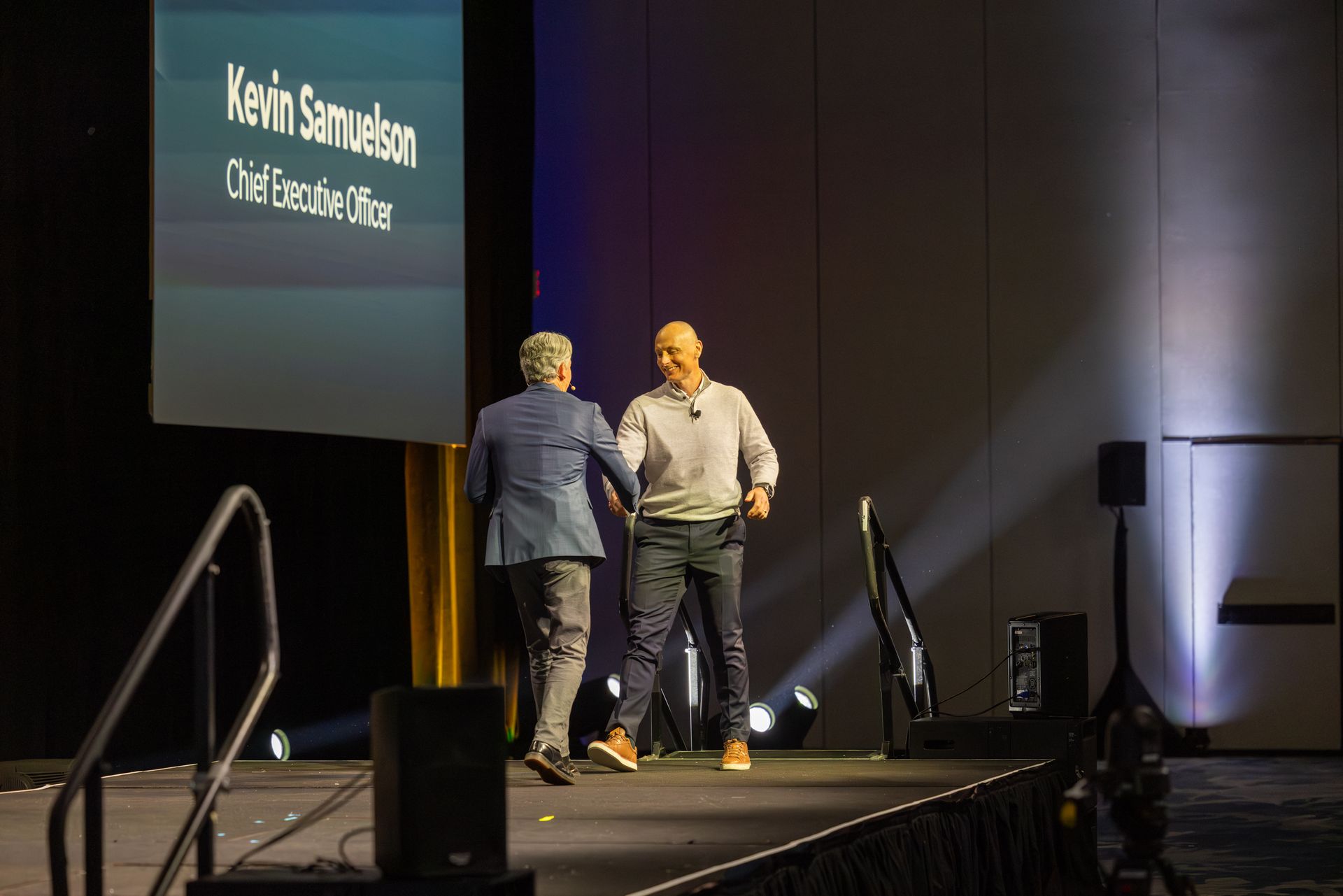 Two men shaking hands on a stage. One in a suit, the other in casual wear, under a screen with text.