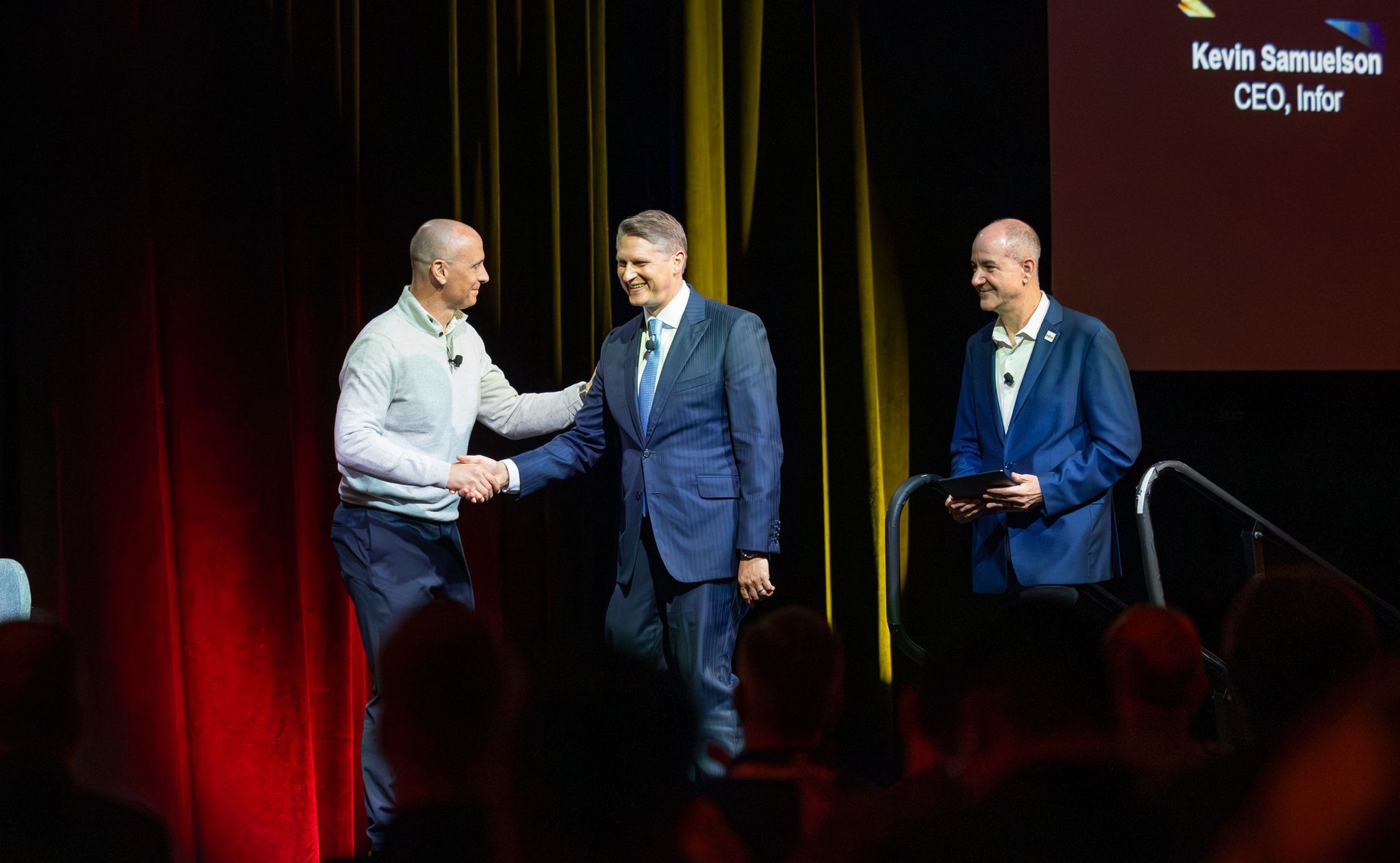Three men on a stage; one shakes hands with another. A screen displays a name and title: Kevin Samuelson, CEO, Ivanti.