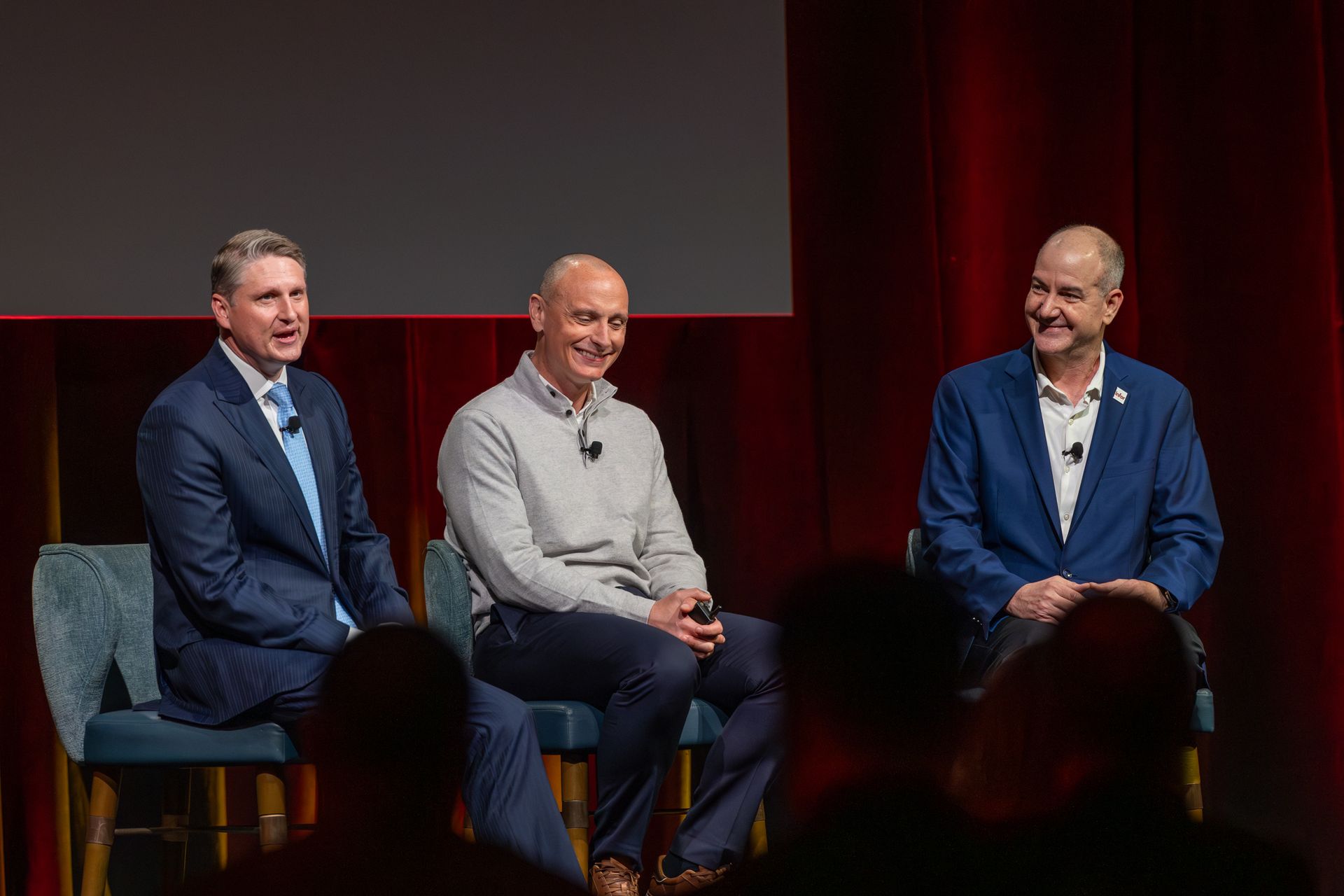 Three men seated on stage, talking. Two are in blue jackets, one in a gray sweater. Red curtains in background.