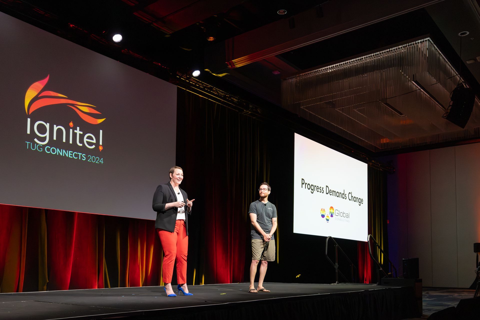 Two people on stage at a conference; one woman in a blazer and red pants speaks, other person stands.