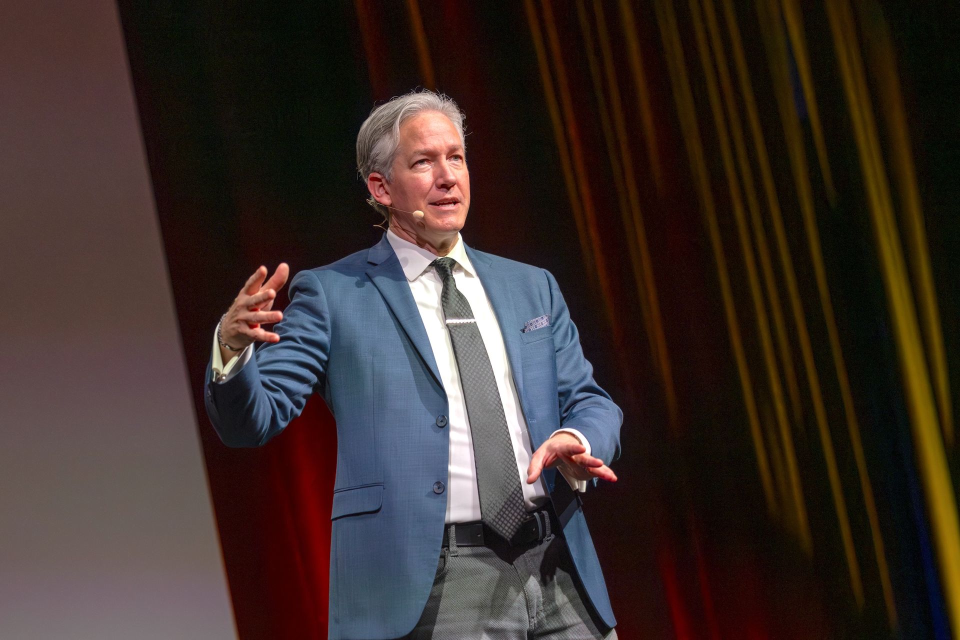 Man in blue suit gestures on a stage with red curtains.