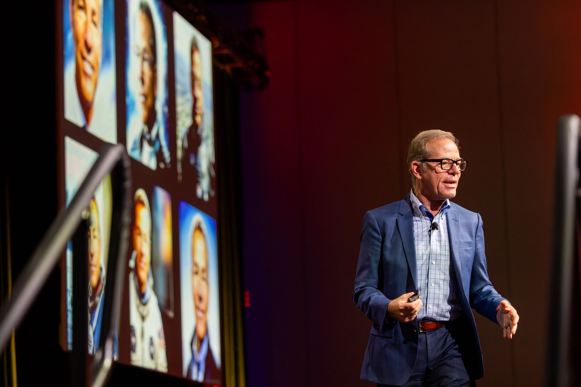 Man in blue suit gestures on stage, images of astronauts displayed on screen behind him.