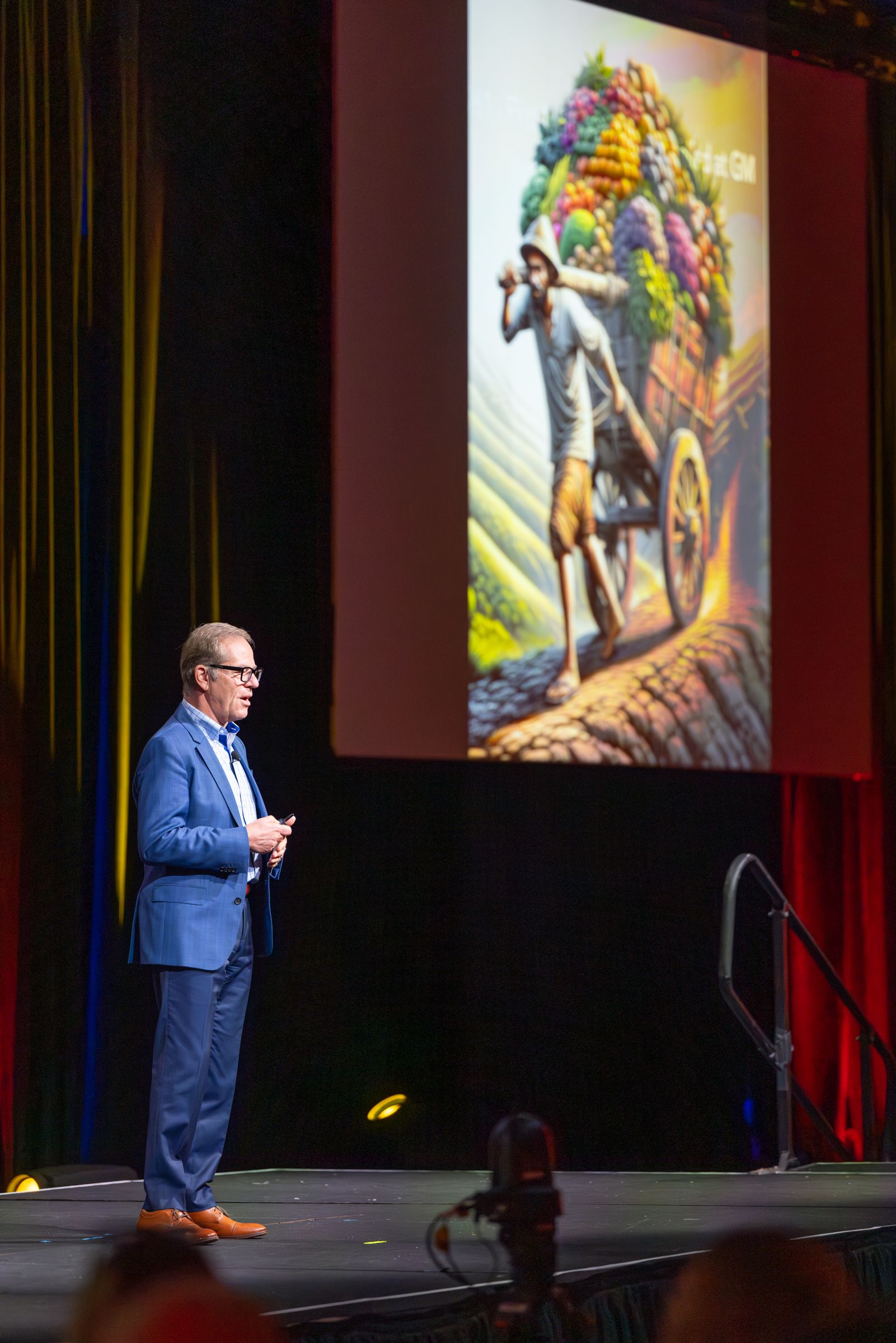 Man in blue suit presents on stage; slide shows person carrying cart of colorful plants.