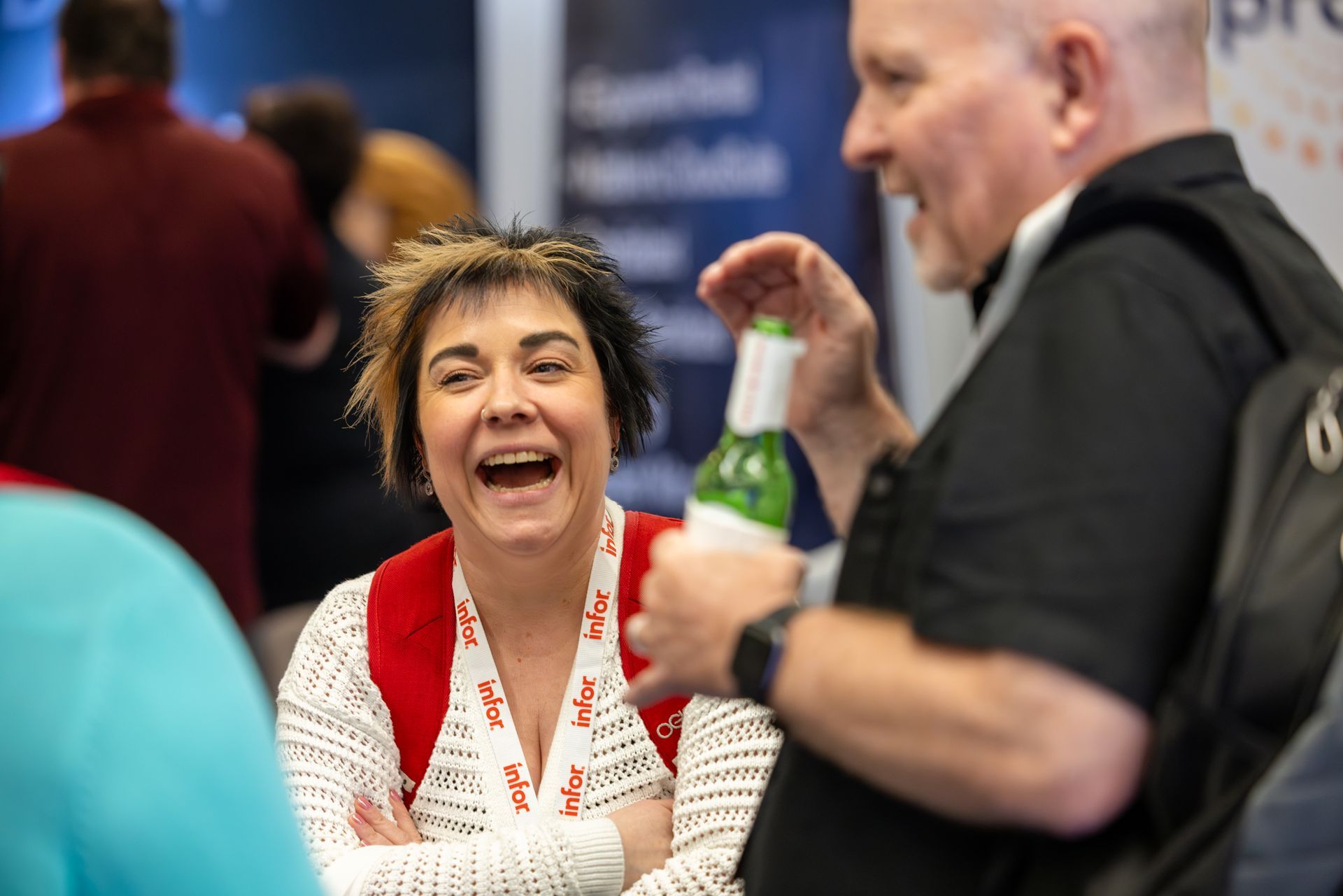Woman laughing, arms crossed, facing a man holding a bottle. Both appear joyful at an event.