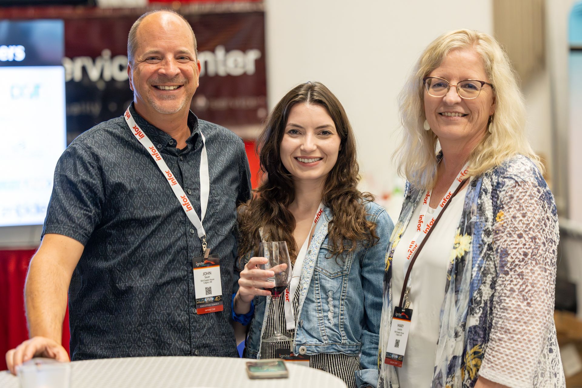 Three people smiling at an event; man in patterned shirt, woman in denim jacket, and woman in floral top.