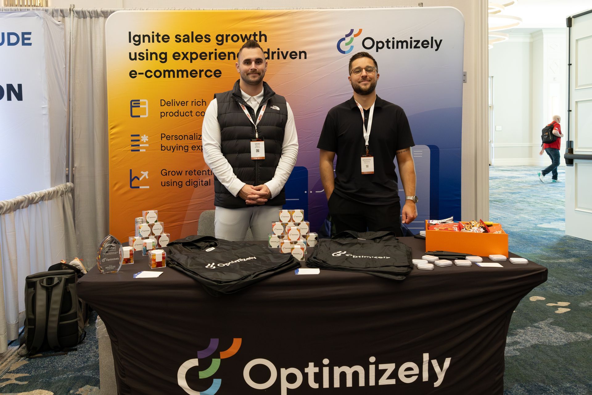Two men at an Optimizely trade show booth, promoting e-commerce. Table with swag and snacks.