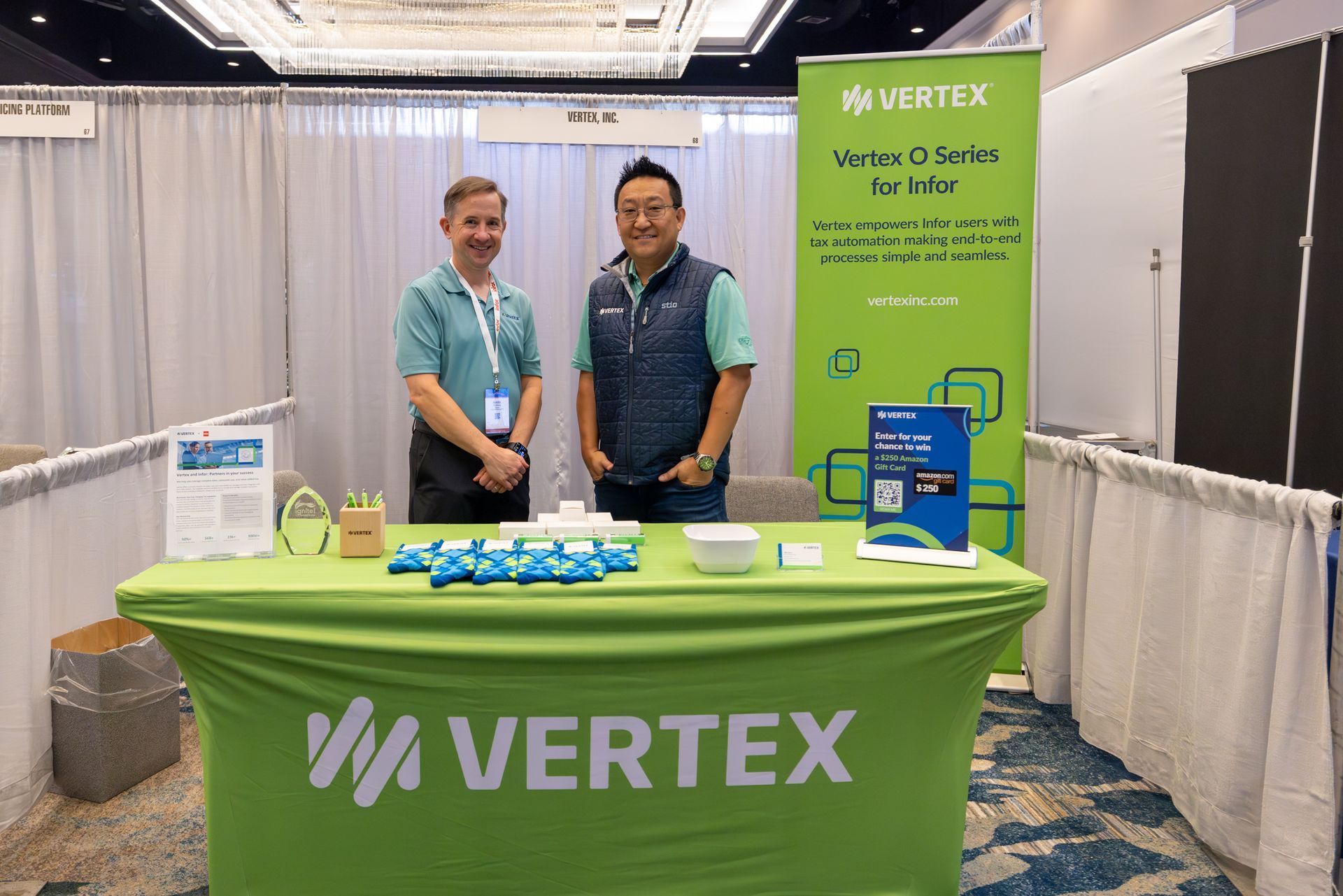 Two men at a trade show booth with a green Vertex banner and table.