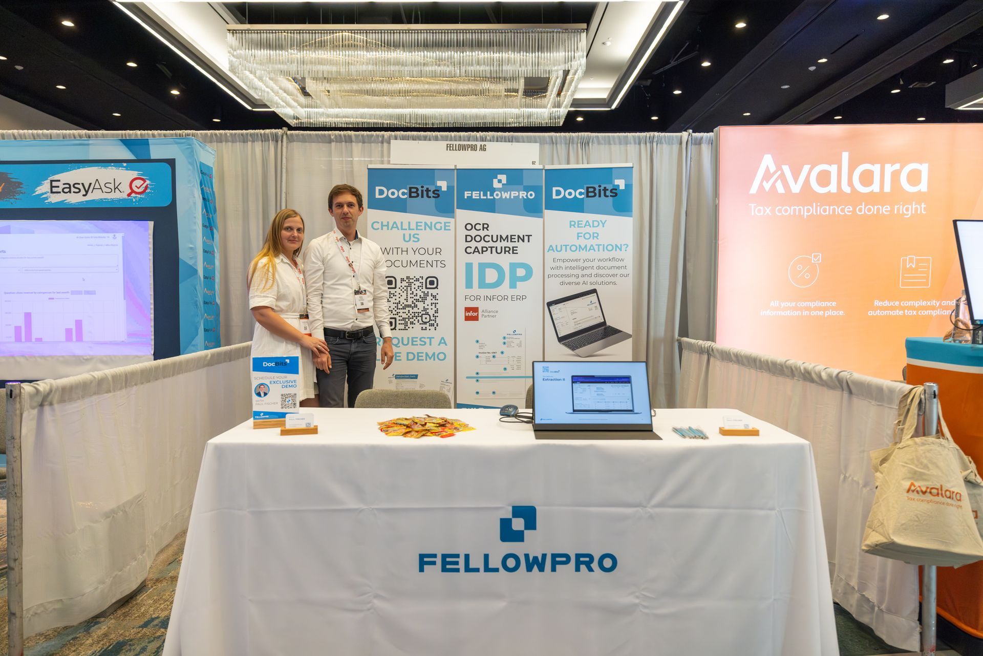 Two people at the FellowPro booth at a conference, with a table, banner, and laptop.