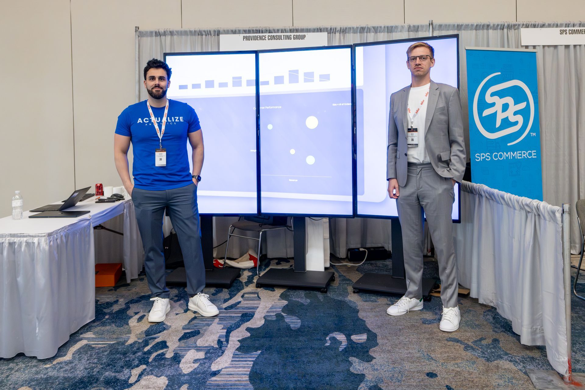 Two men stand near three display screens at an event booth. One wears a blue shirt, the other a grey suit.