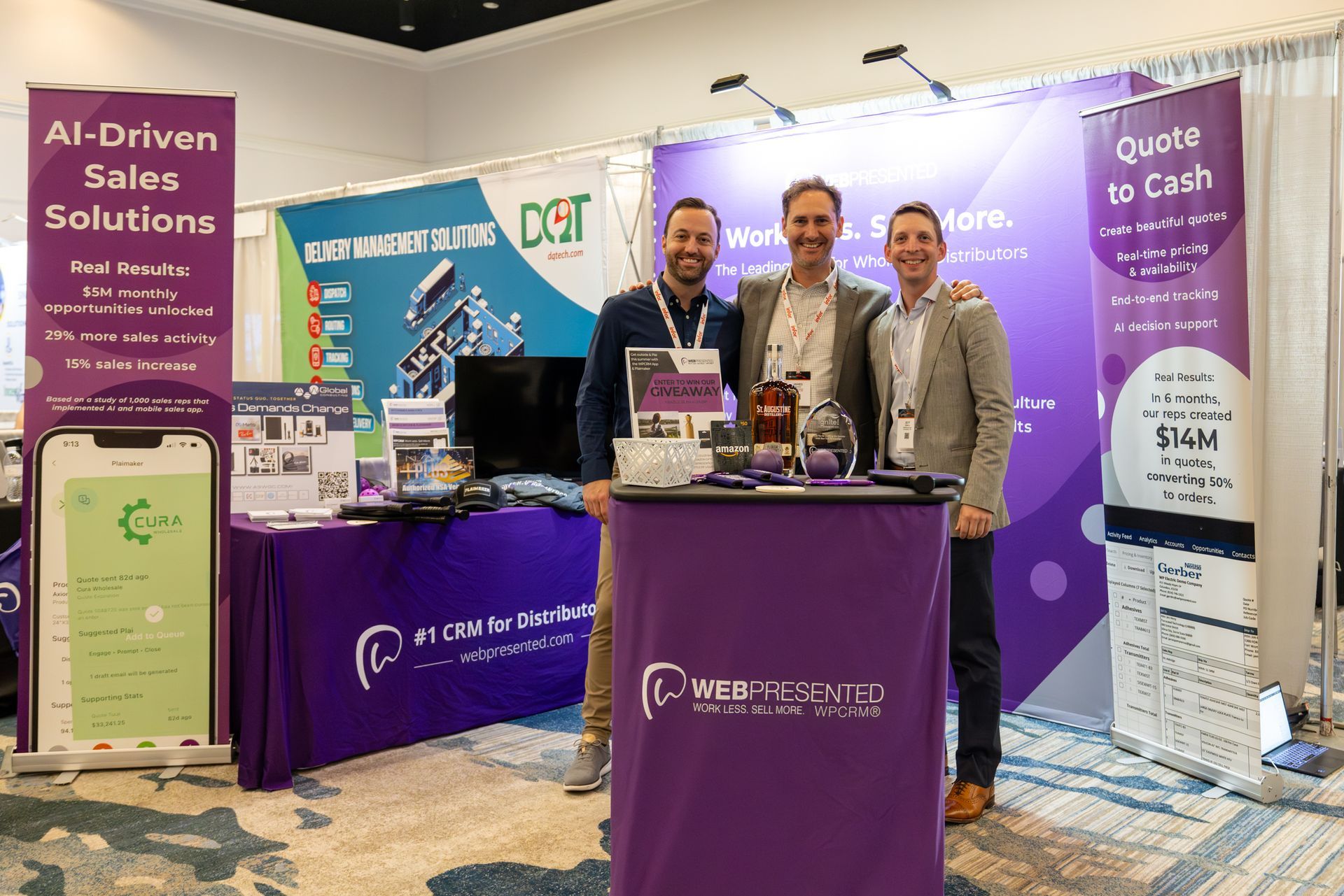 Three men at a trade show booth, purple and white signage, smiling, standing behind a table, indoor setting.