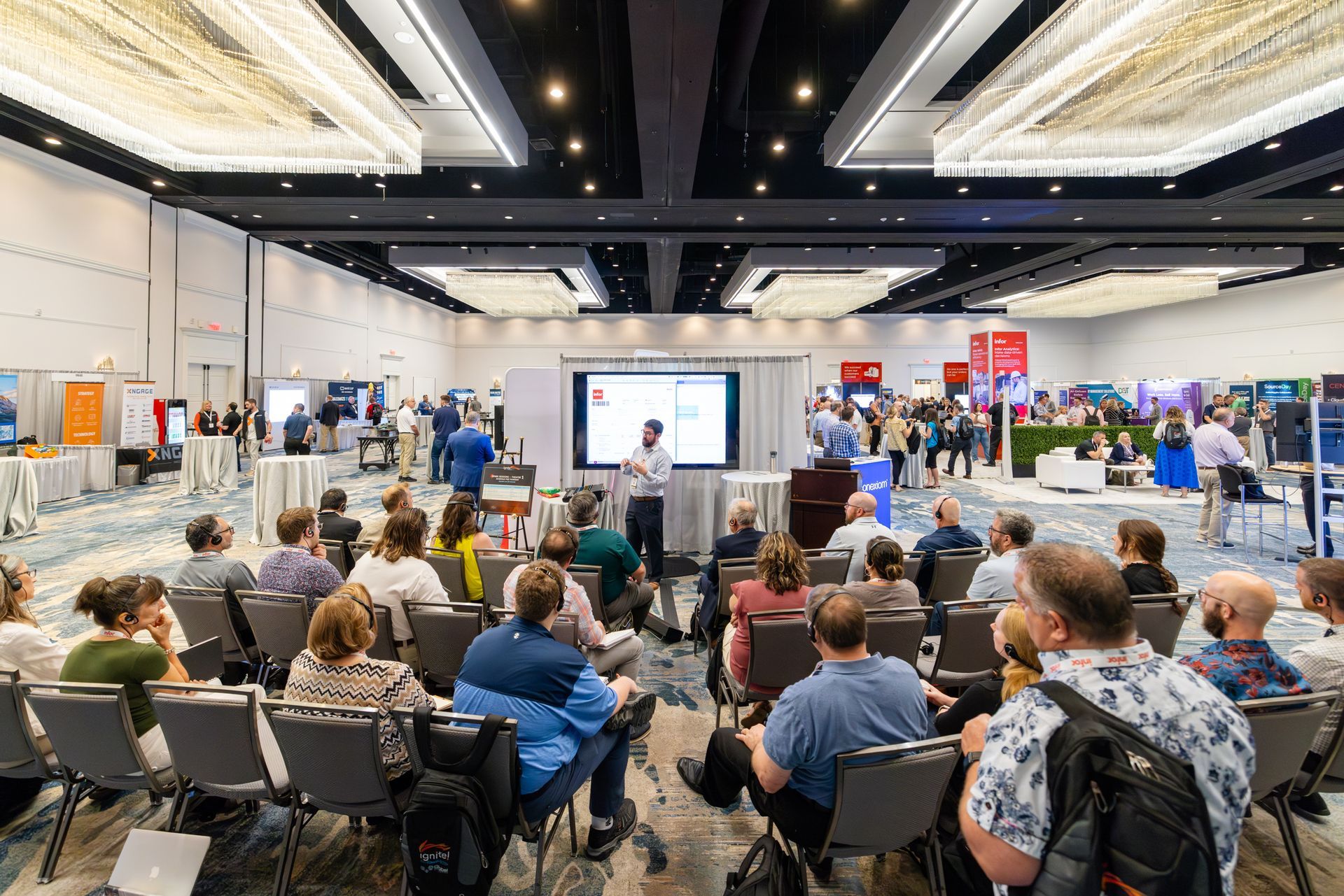 Audience at a conference presentation; speaker stands near a screen, many attendees in a large room.