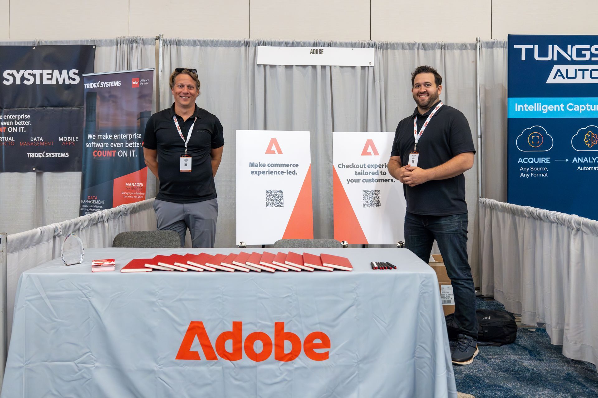Two men stand behind an Adobe booth at a conference. They are smiling, and promotional materials are visible.