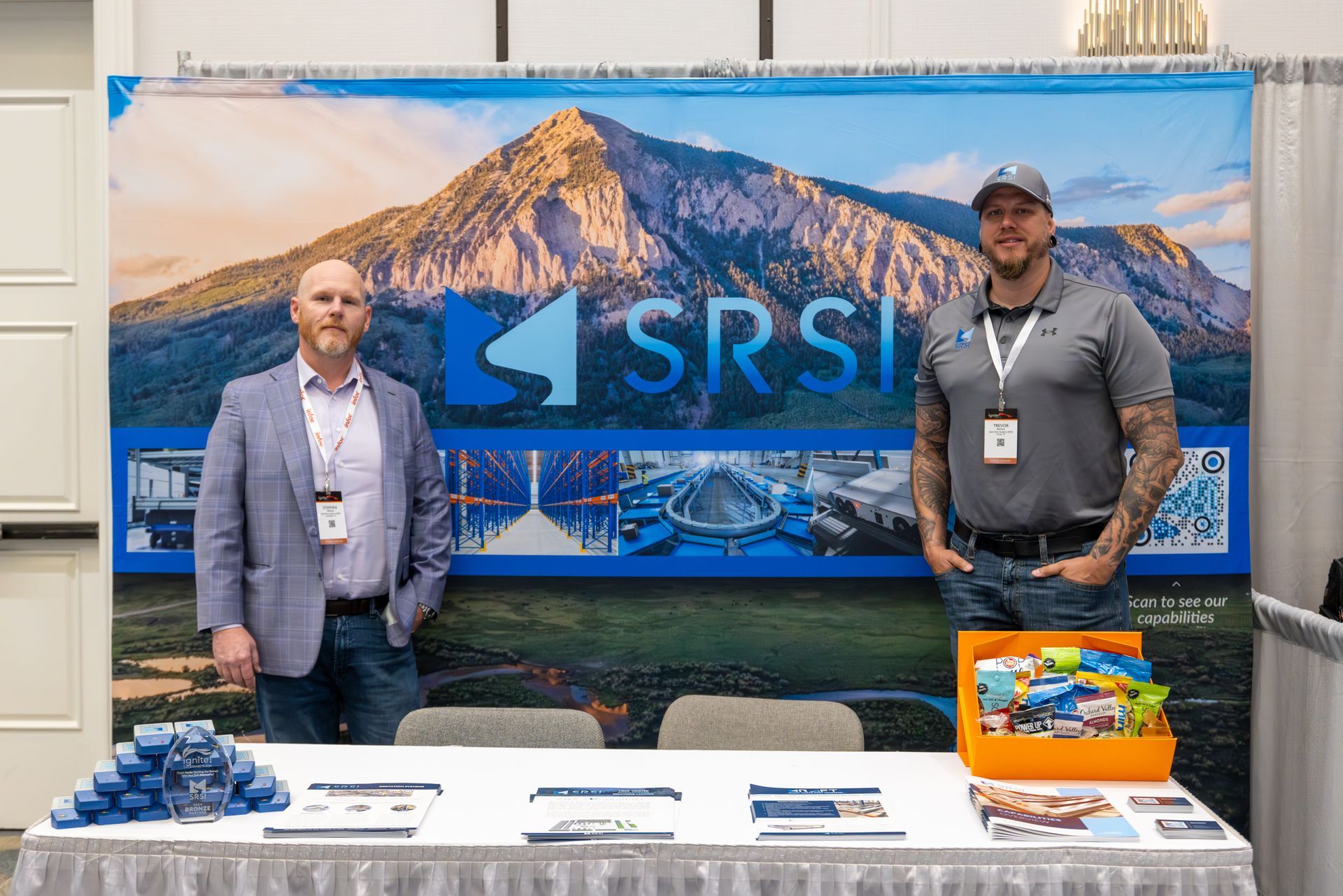 Two men at a booth with a mountain backdrop and SRSI logo. They're at a conference or trade show.