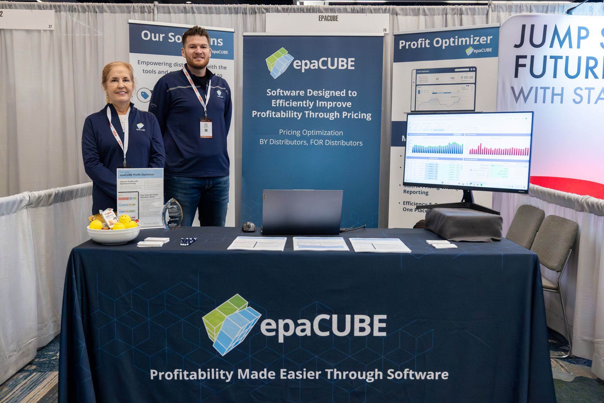 Two people at a trade show booth for epaCUBE. Signage, laptop, graphs, and a table with materials are present.