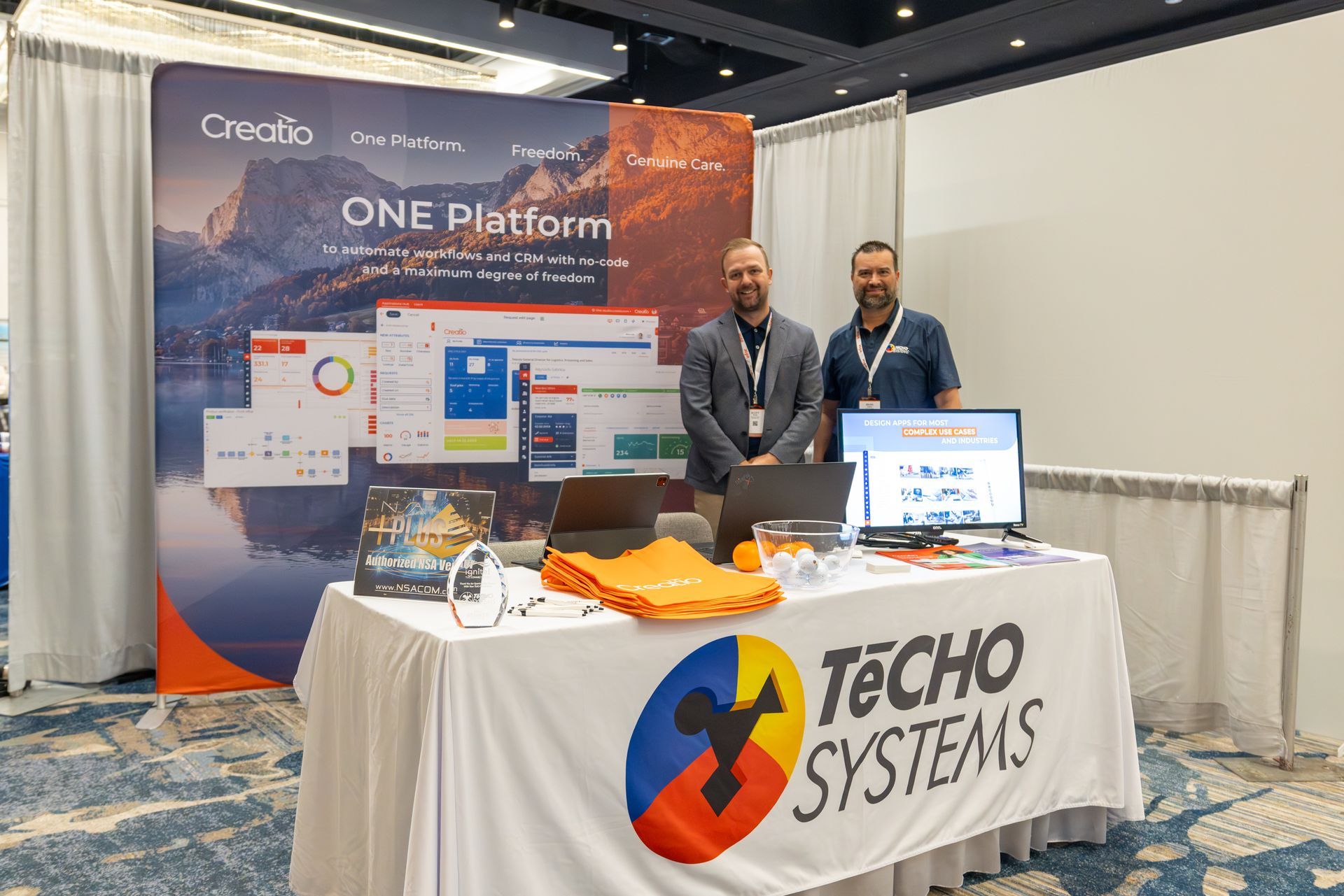Two men stand at a trade show booth for Techo Systems. Booth displays product visuals and branding.