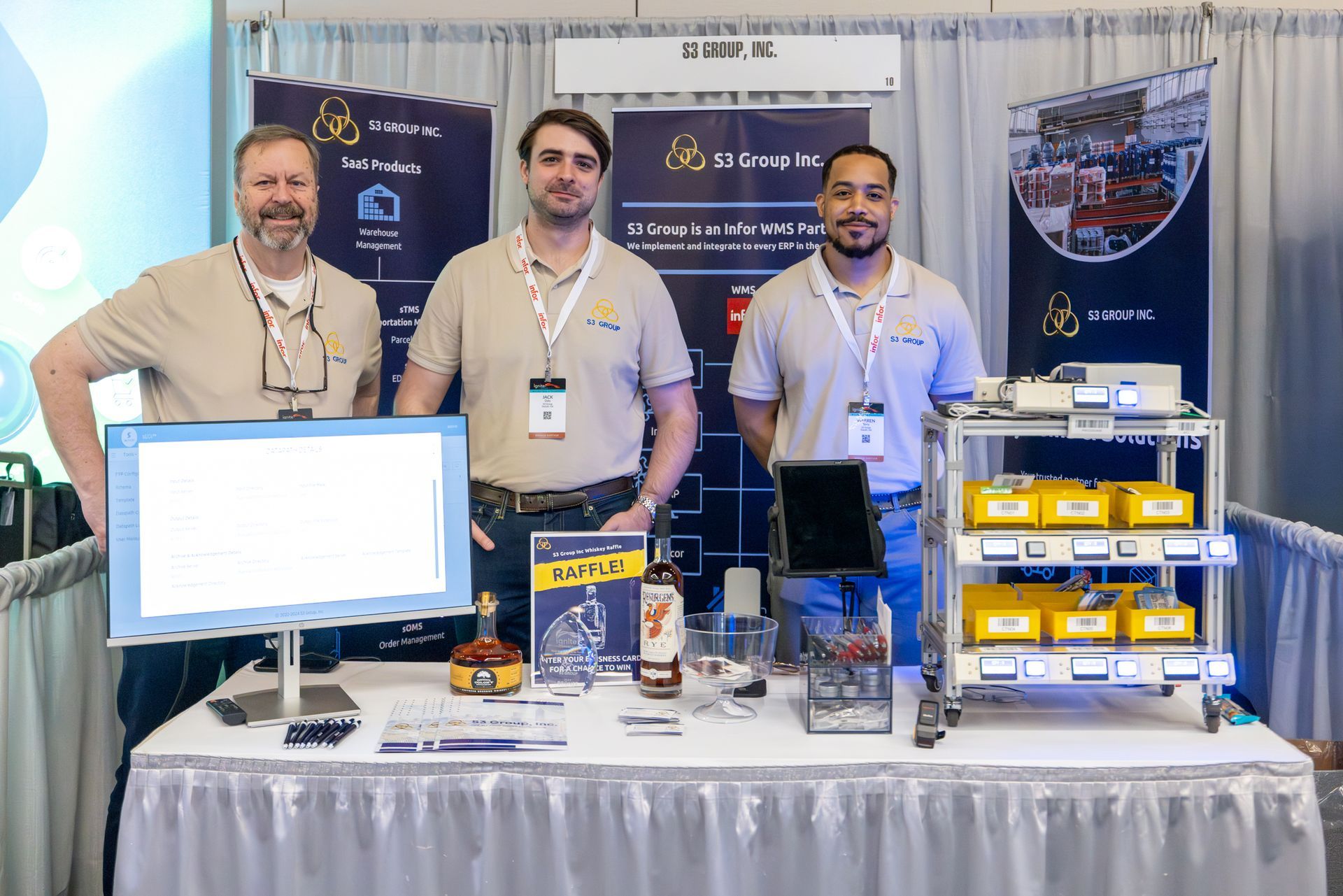 Three men stand behind a booth at a trade show. They are in front of a table with equipment and display boards.