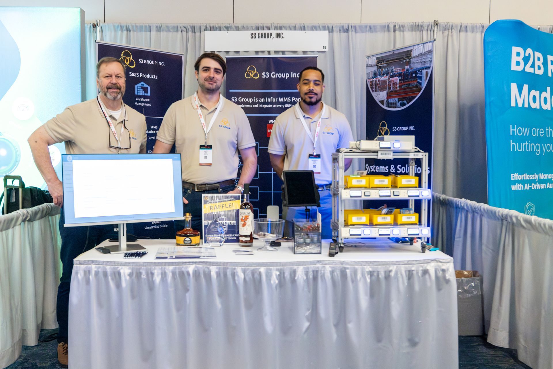 Three men at a trade show booth, behind a table with tech displays. Logos and banners visible.