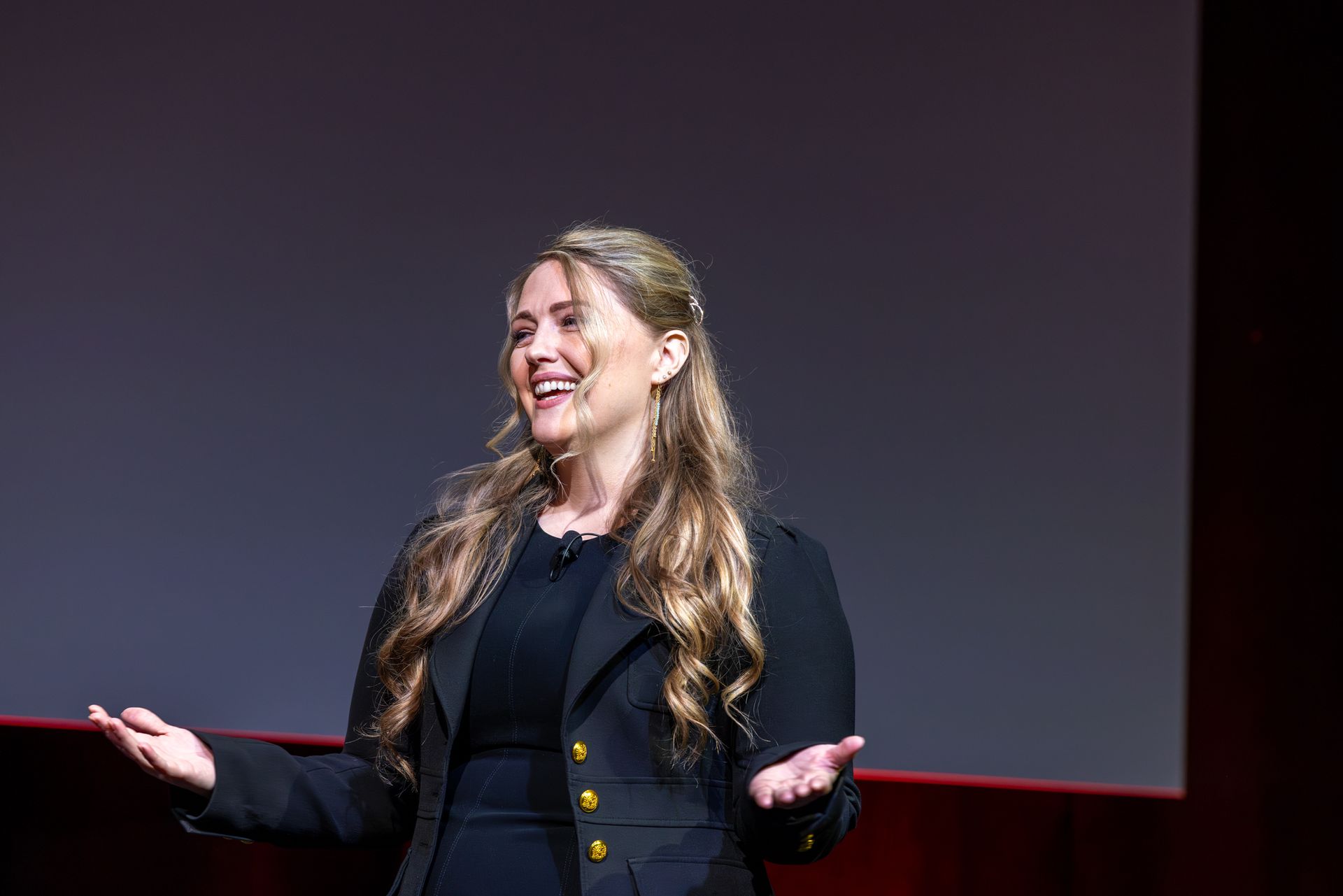 Woman on stage with arms open, smiling. Wearing a black blazer, facing a blank screen.