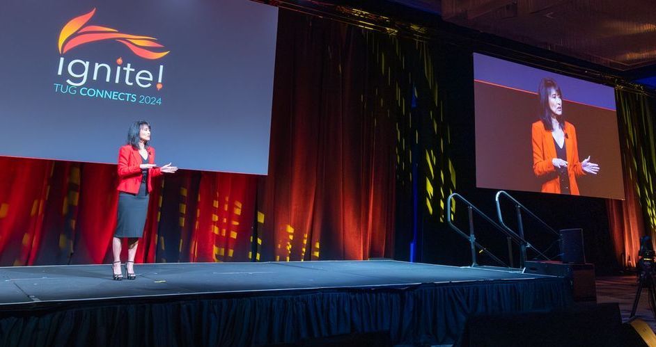 Woman in red blazer speaking on stage at a conference, with a screen displaying 