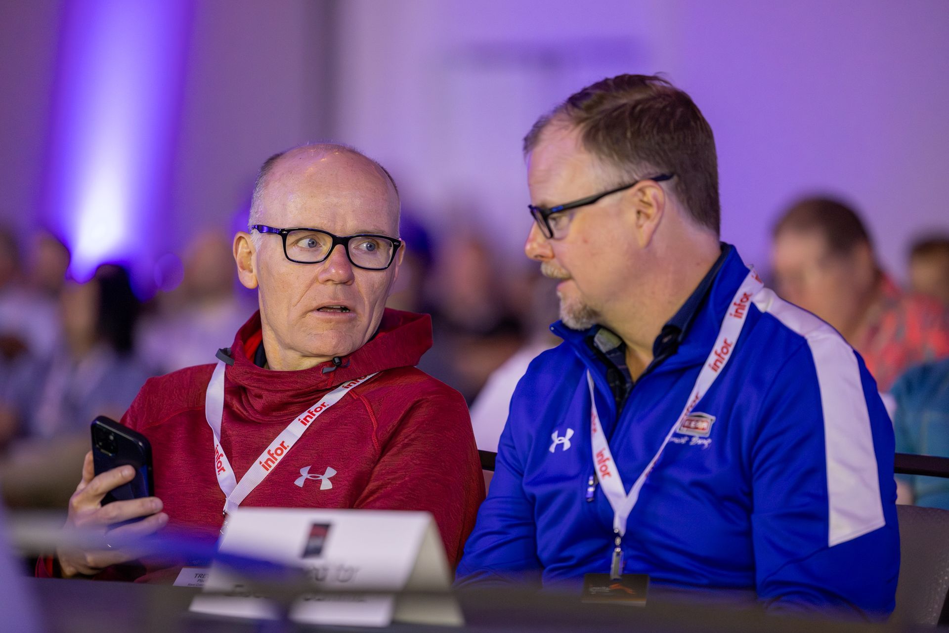 Two men in a conference setting; one holds a phone, the other speaks. Both wear glasses and lanyards.