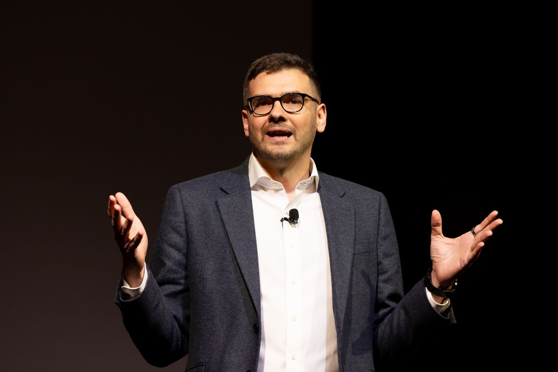 Man in blazer and glasses speaks with hands raised on stage, dark background.
