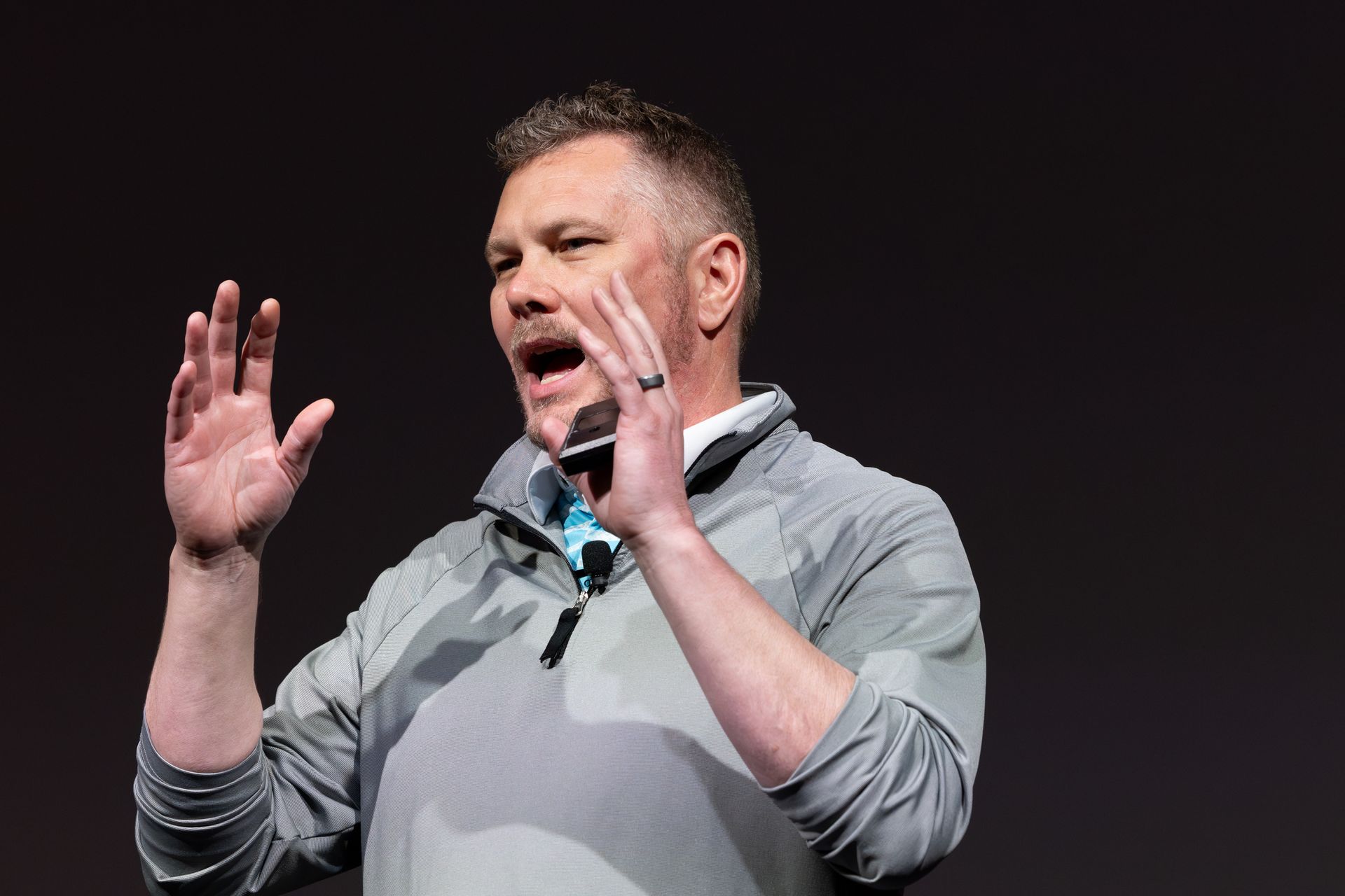 Man speaking with hands raised, wearing a gray jacket, open mouth, microphone, dark background.