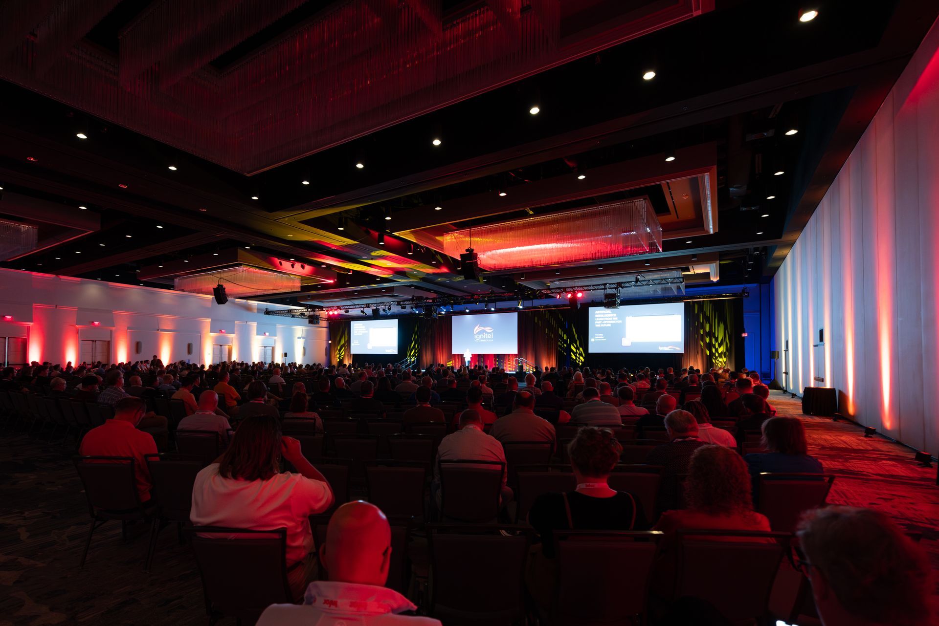 Large conference hall filled with people attending a presentation; two screens display content, red and white lighting.